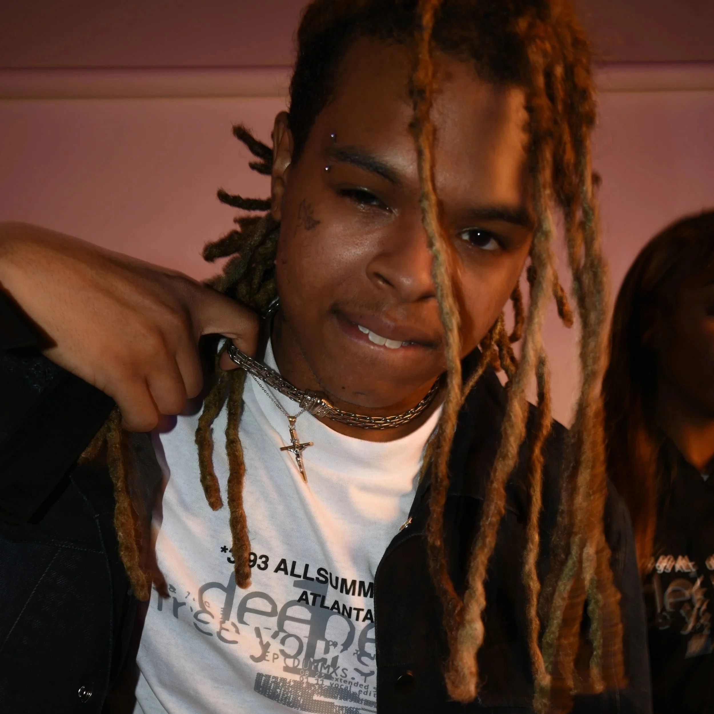 Close-up of a person's face with dreadlocks, multiple facial piercings, and facial tattoos, wearing a white shirt and layered necklaces, smiling at the camera.