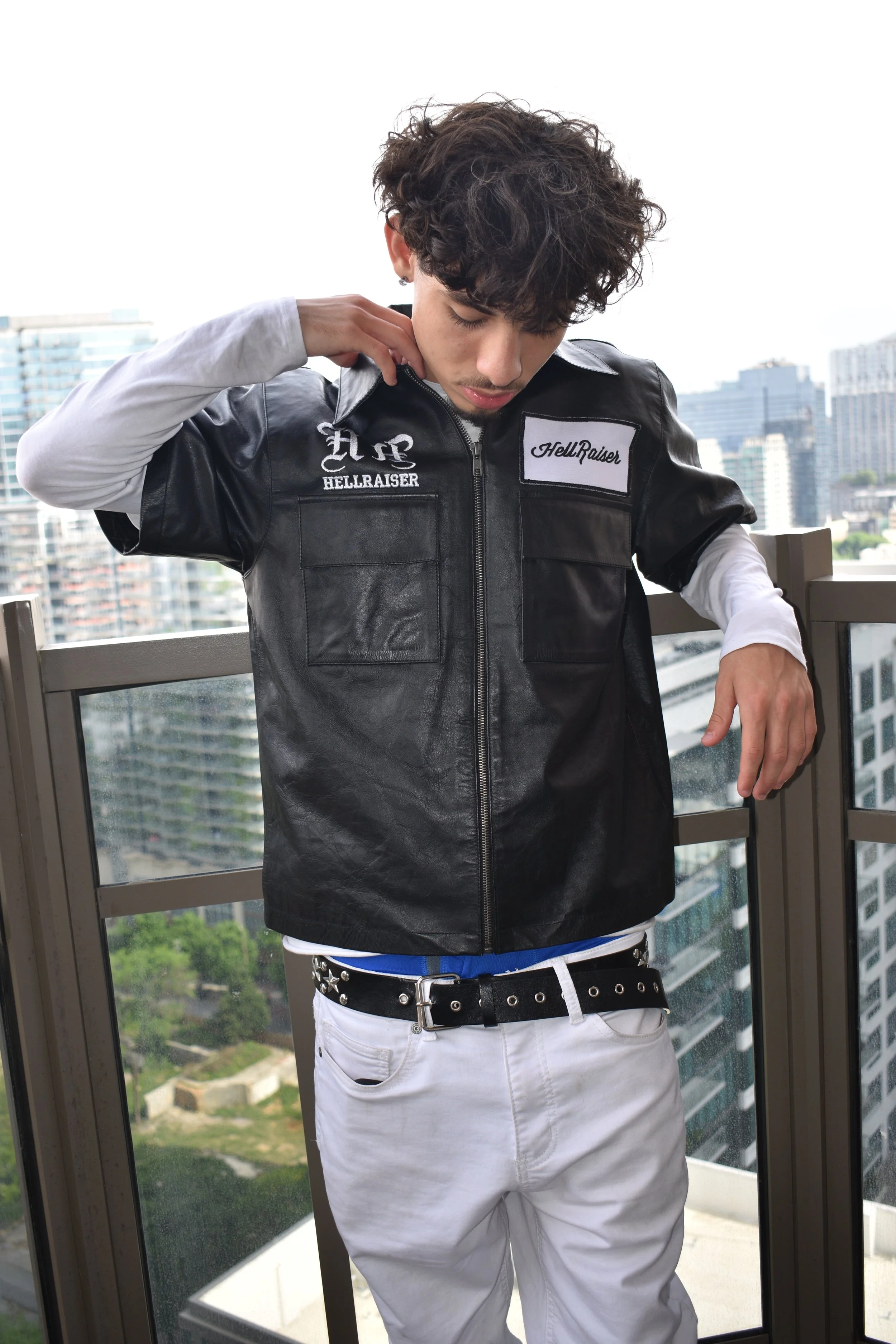 A young man looking down while adjusting his black leather vest, standing on a balcony with a cityscape in the background.