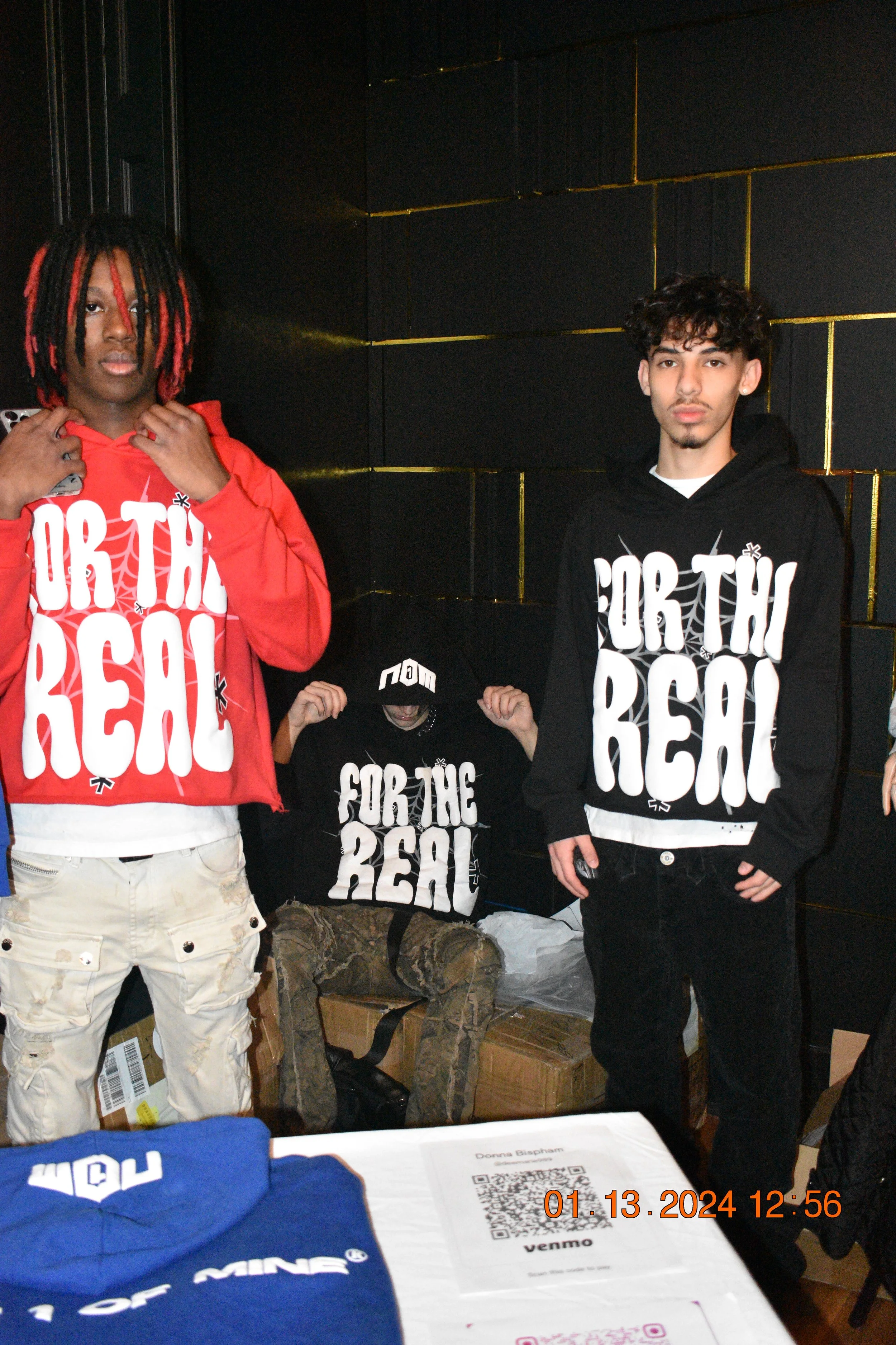 Three young men wearing hoodies with the phrase 'FOR THE REAL' printed on them, standing and sitting in front of a black wall with gold accents, in an indoor setting.
