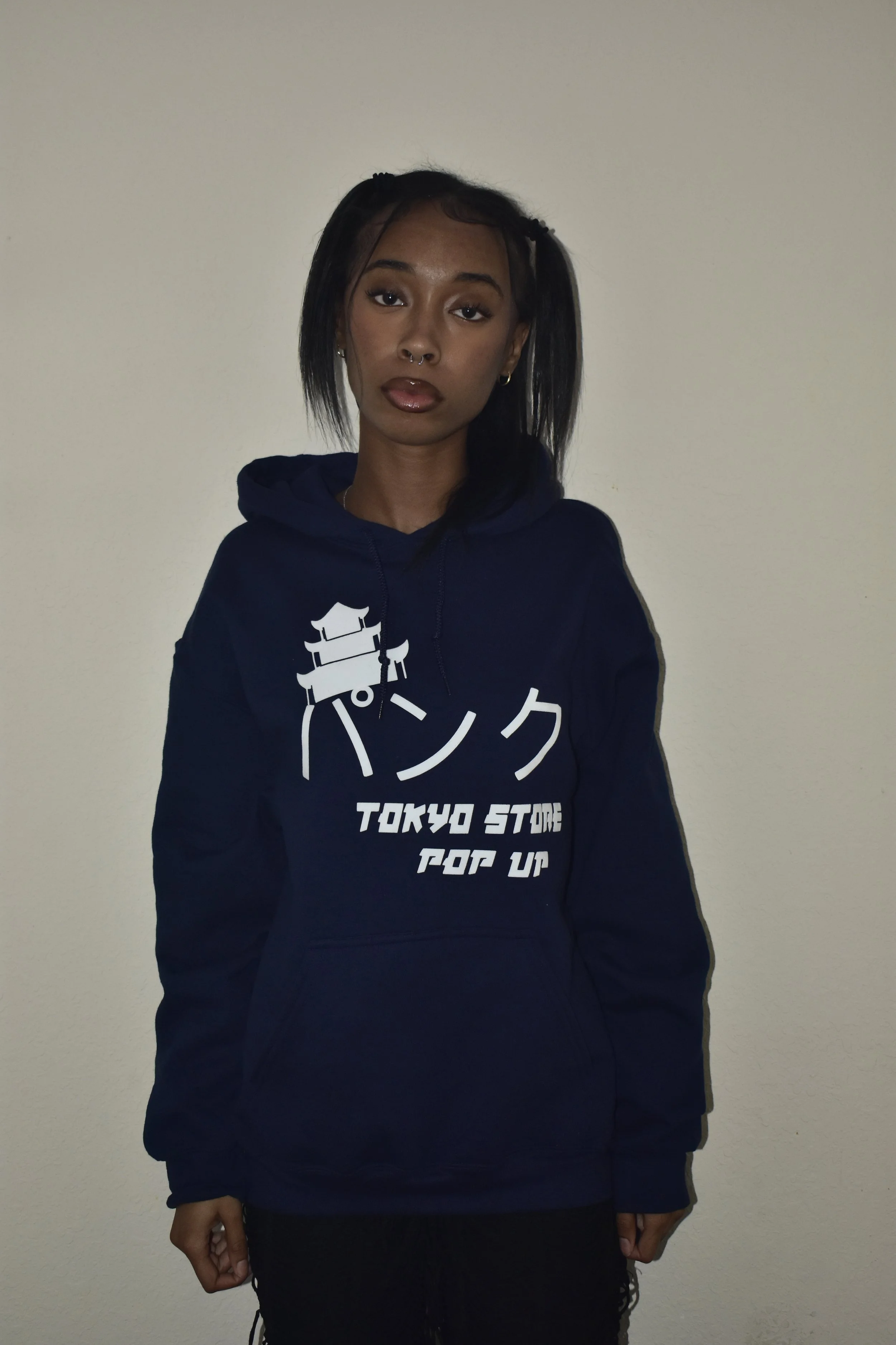 A young woman with dark hair, wearing a navy hoodie with white Japanese characters and English text, standing against a plain wall.