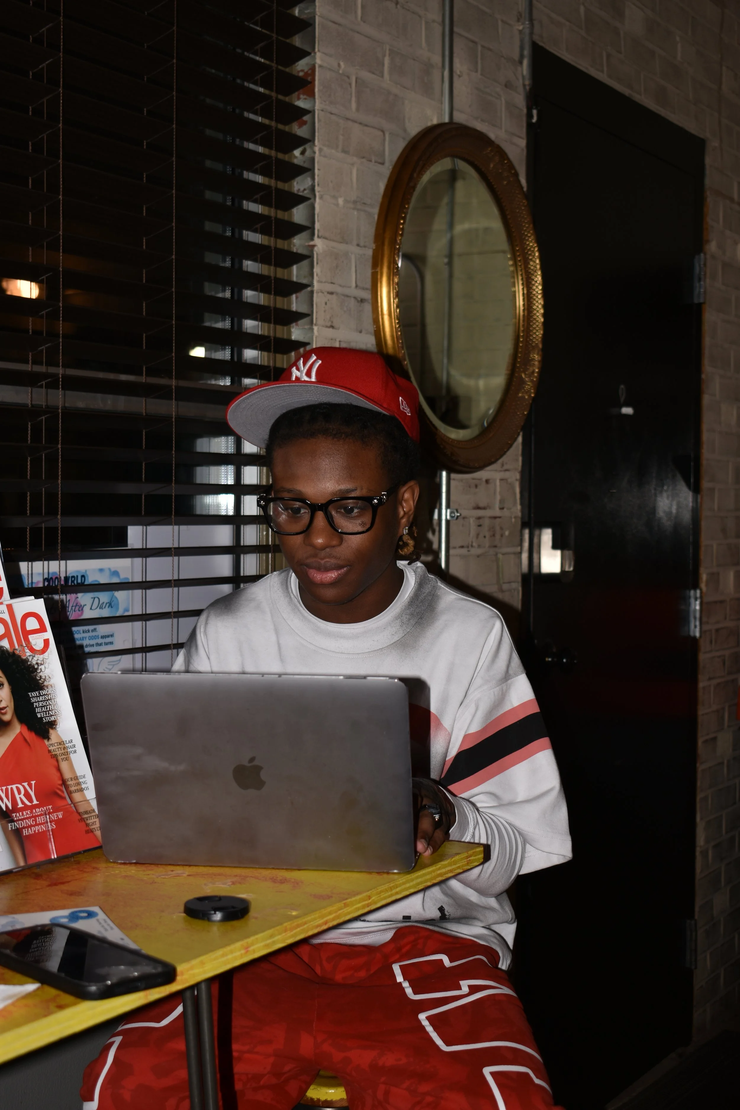 A person with glasses and a red cap sitting at a table, working on a laptop in a dimly lit cafe or restaurant. There is a magazine on the table and a smartphone next to it.