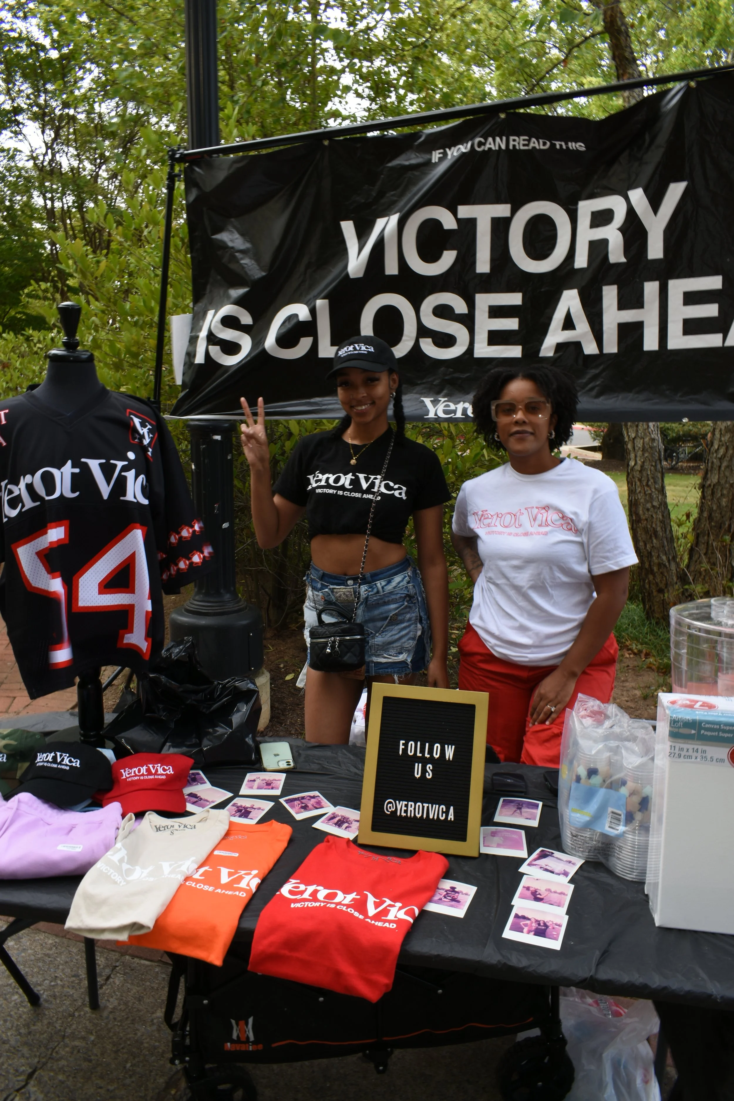 Two women standing behind a table with merchandise, including T-shirts, caps, and photos, at an outdoor event. One woman is making a peace sign and the other is standing with her hand on her hip. A large banner behind them reads 'VICTORY IS CLOSE AHE