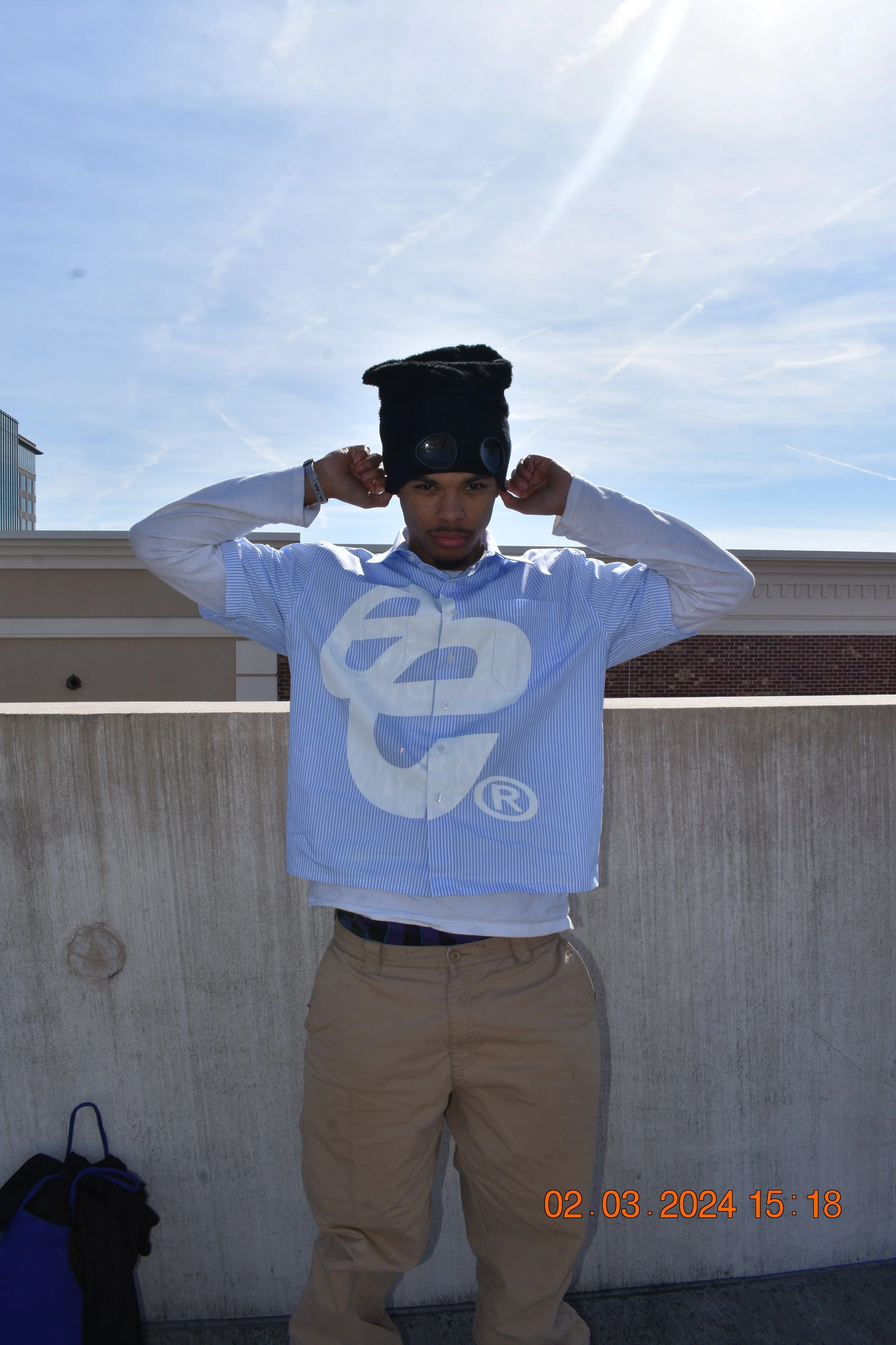 A young man wearing a black beanie and a blue and white striped shirt with a large logo, standing outdoors against a concrete wall and blue sky with wispy clouds. He is adjusting his beanie with both hands and looking directly at the camera.