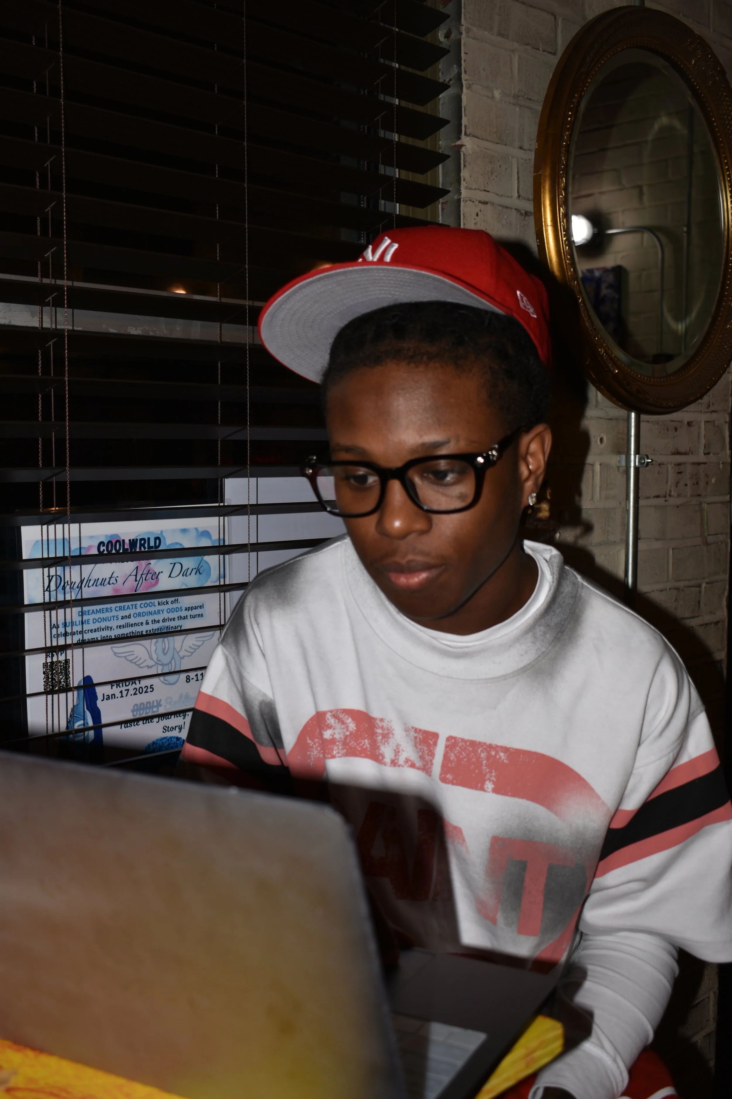 A young person with glasses and earrings, wearing a red and white cap and a white sweatshirt with red and black design, working on a laptop in a dimly lit room with blinds, a mirror, and a poster in the background.
