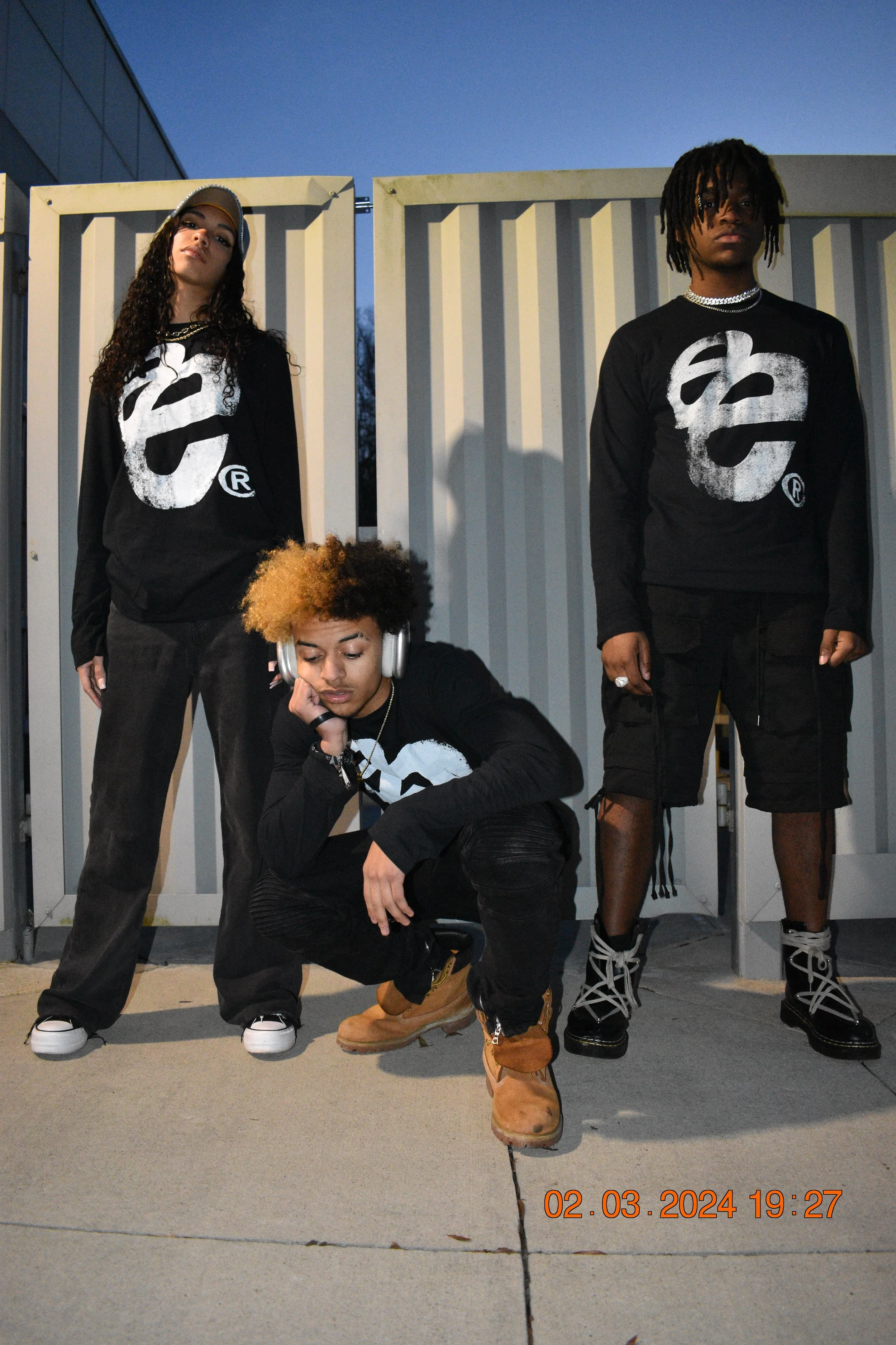 Three young people posing outdoors during sunset, wearing matching black shirts with a white logo, one crouching with headphones, two standing against a white fence with a city building in the background.