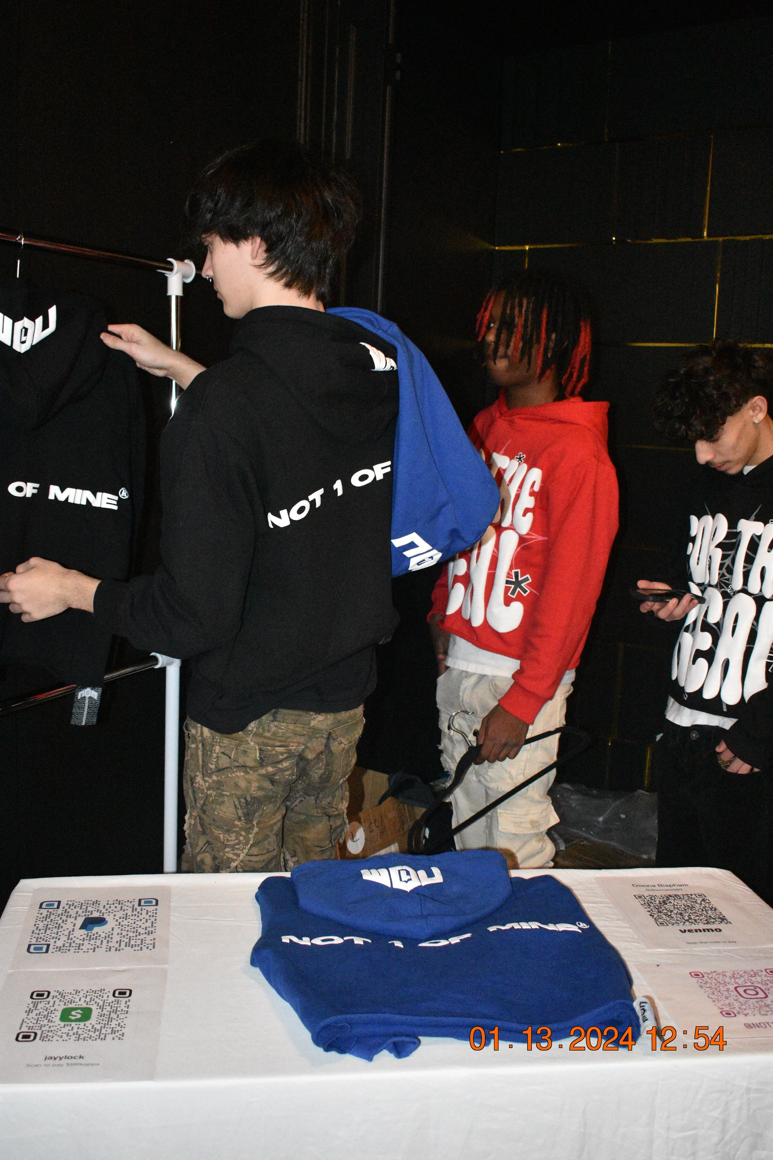 Three young men standing at a table with blue hoodies and QR codes on it, in a dimly lit room, possibly at an event or store, with one looking at their phone and another holding a shopping cart.