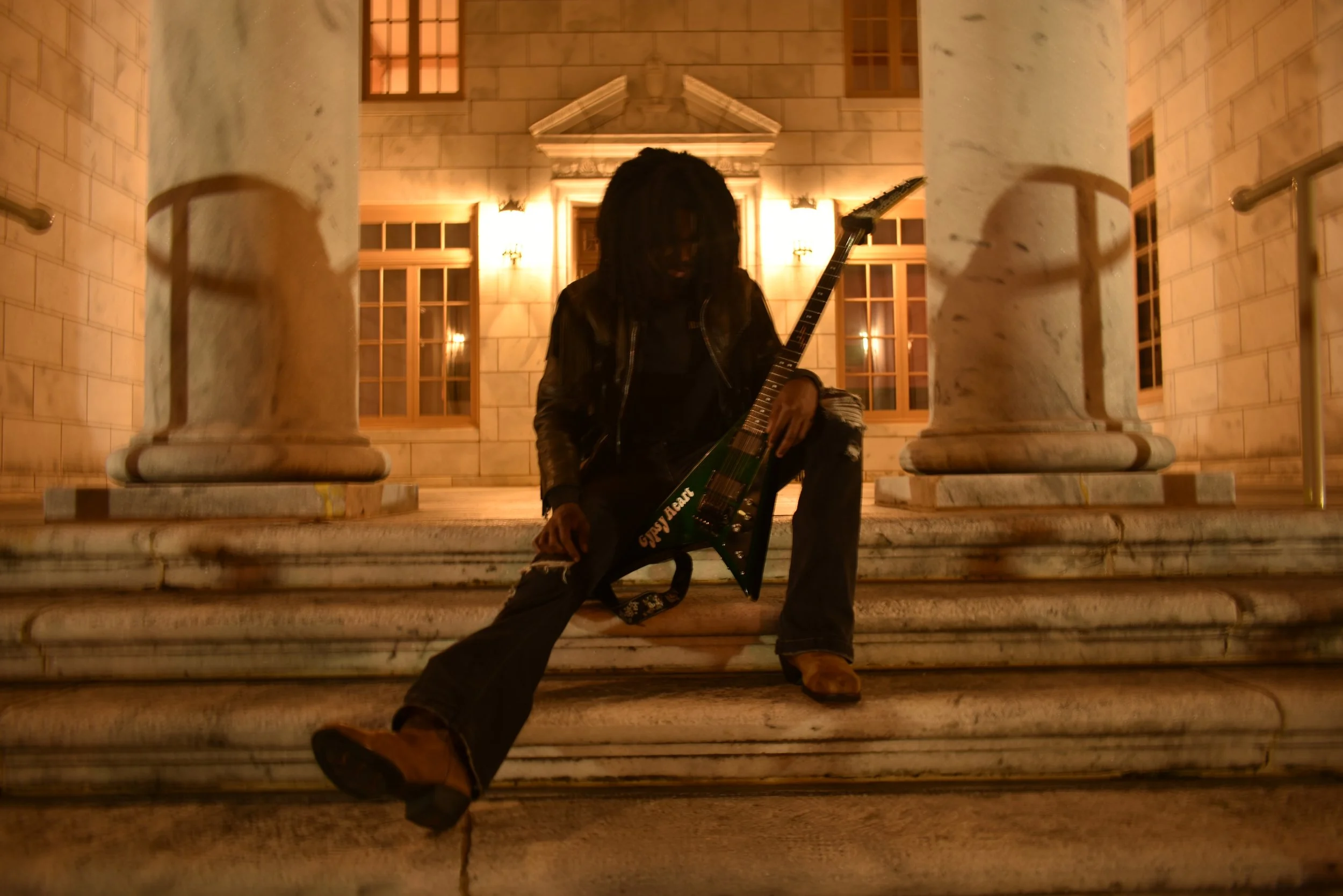 A person with dreadlocks sitting on steps of a building at night, holding an electric guitar, wearing a black leather jacket and ripped jeans, with a classical architectural background.