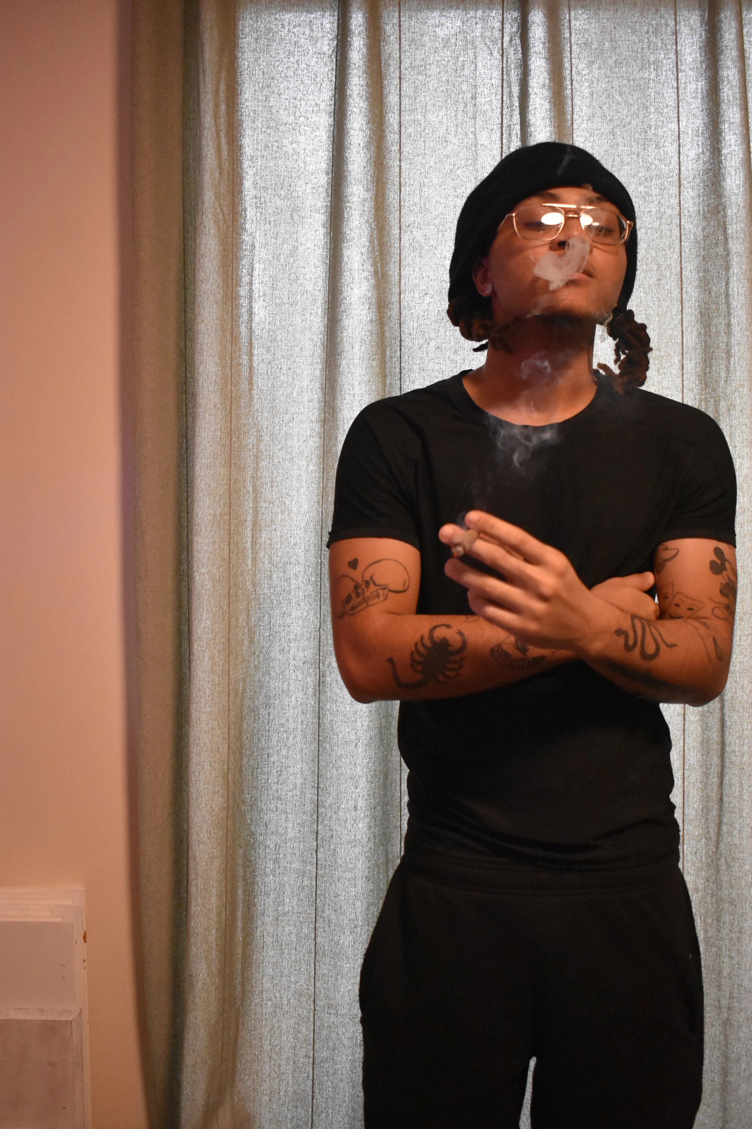 A person stands in front of a curtain, wearing glasses, a black headwrap, and a black t-shirt. They are holding a lit cigarette or joint, producing smoke, and have tattoos on their arms.