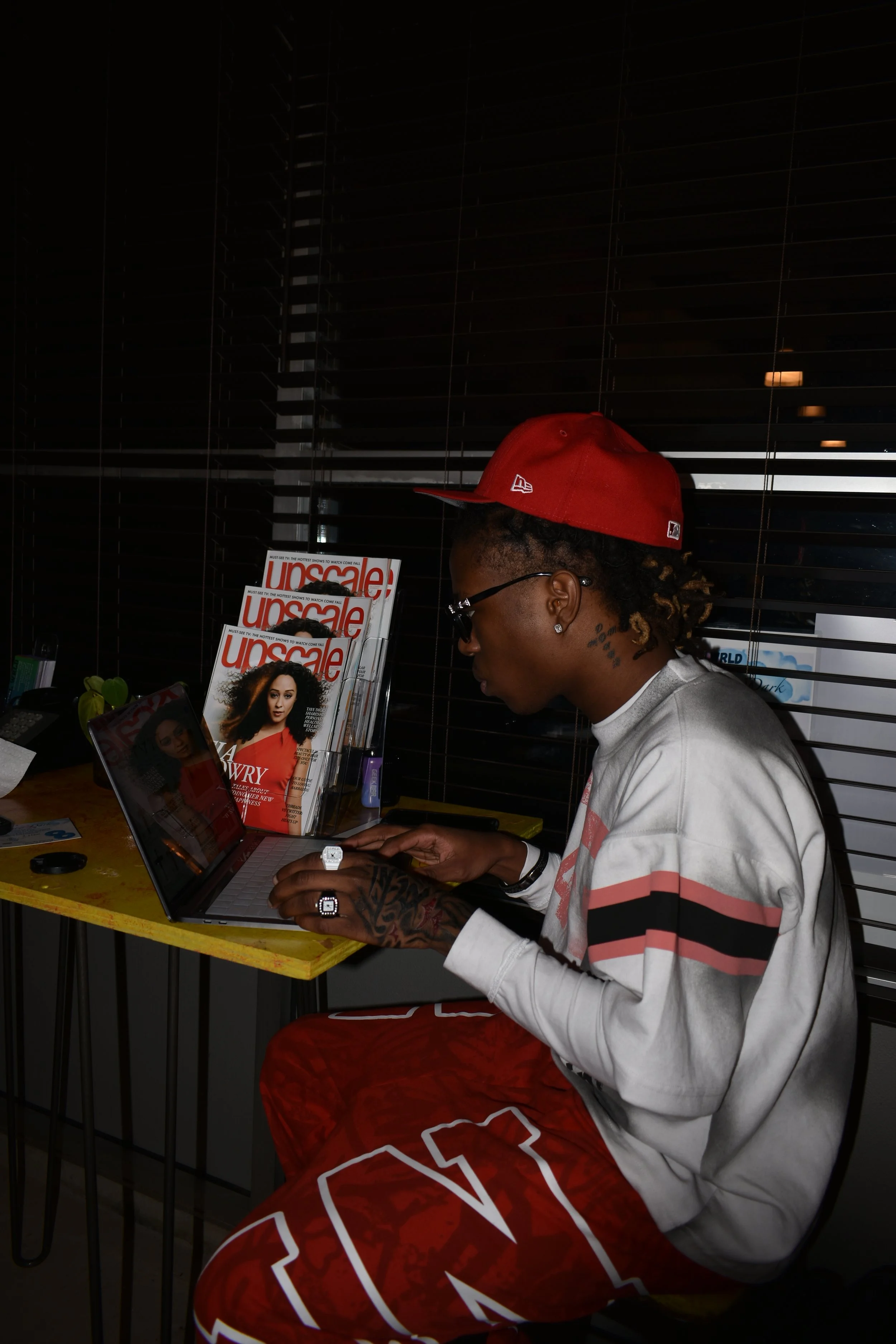 A young man with tattoos and glasses working on a laptop at a yellow table with magazines labeled 'Unsacle' nearby, wearing a red cap, grey sweatshirt, and red pants.