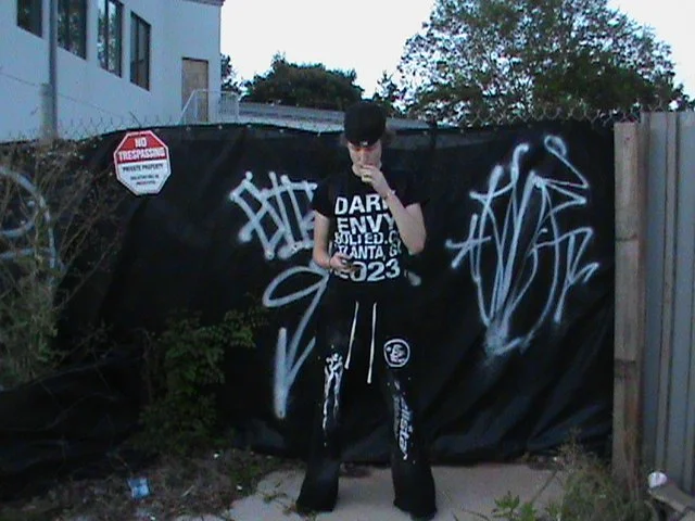 Person with dark hair wearing a black T-shirt with white text, black pants with white designs, and a black cap, standing in an outdoor area with graffiti-covered black fence behind them.