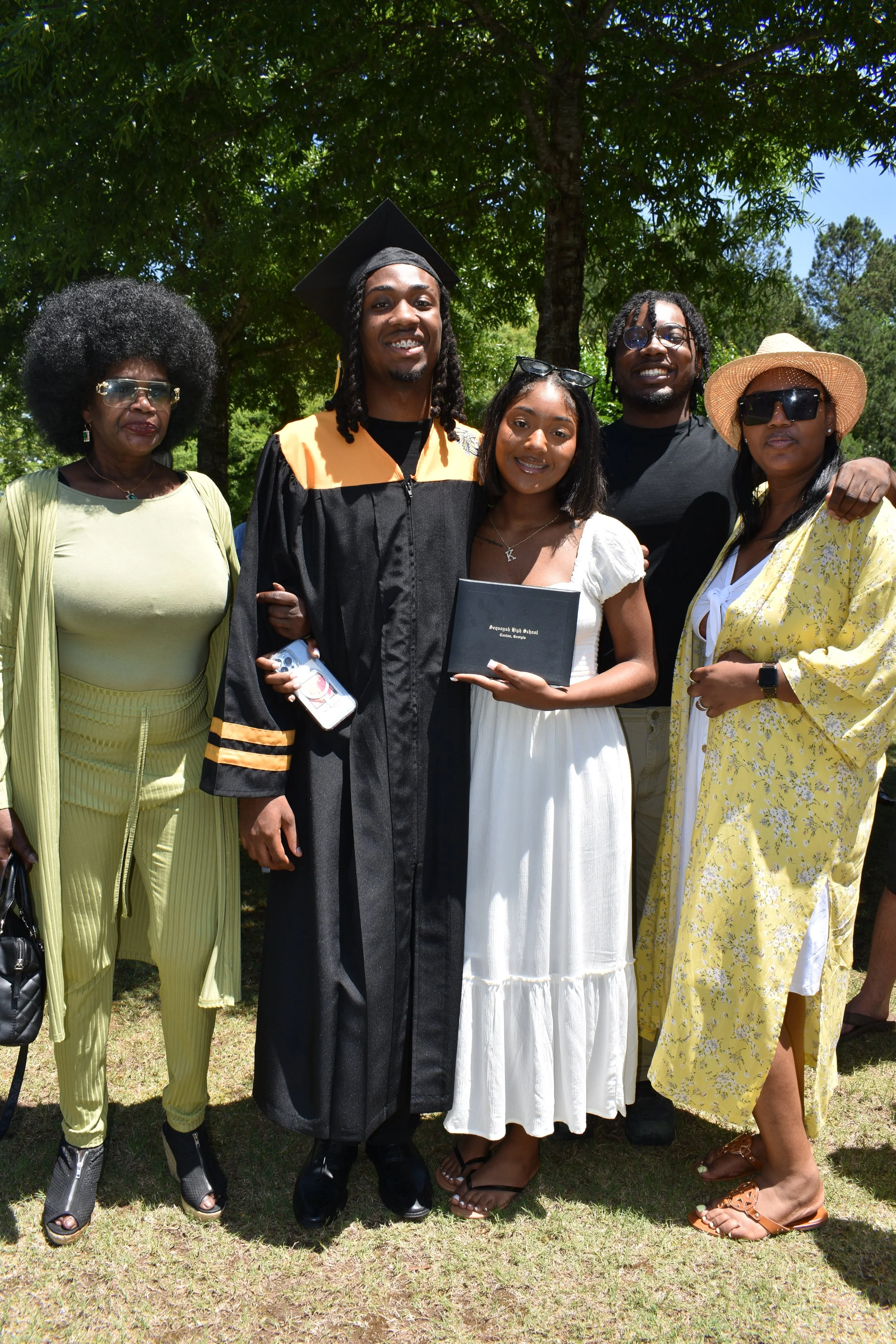 Graduation celebration outdoors with a person in a black cap and gown, holding a diploma, surrounded by family and friends.