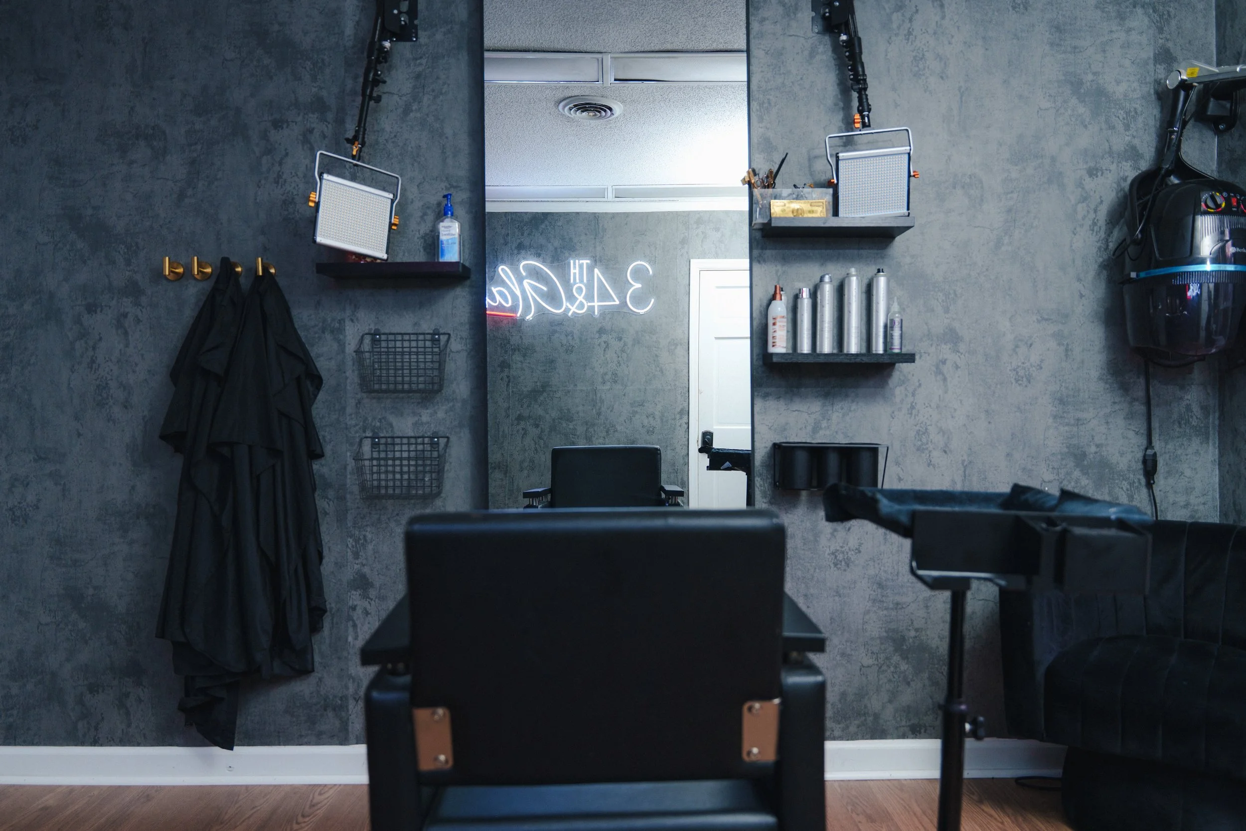 A modern hair salon or barbershop with a large mirror, black chairs, and shelves with styling products. The space has a gray textured wall, a black couch, and various hairdressing tools and containers. Neon signage is partially visible in the reflection.