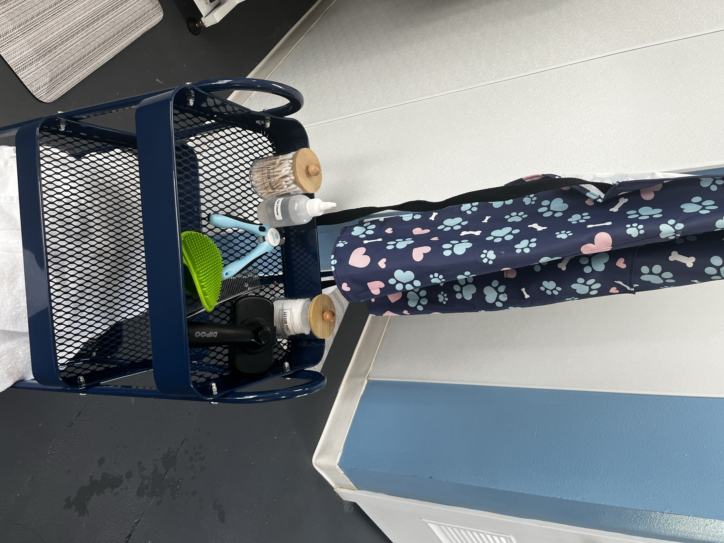 A navy blue cart with a mesh design holding various hair and grooming supplies including a green hairbrush, a black electric shaver, a blue scalp massager, a bottle of liquid soap, and a small container of cotton swabs, positioned against a wall in a