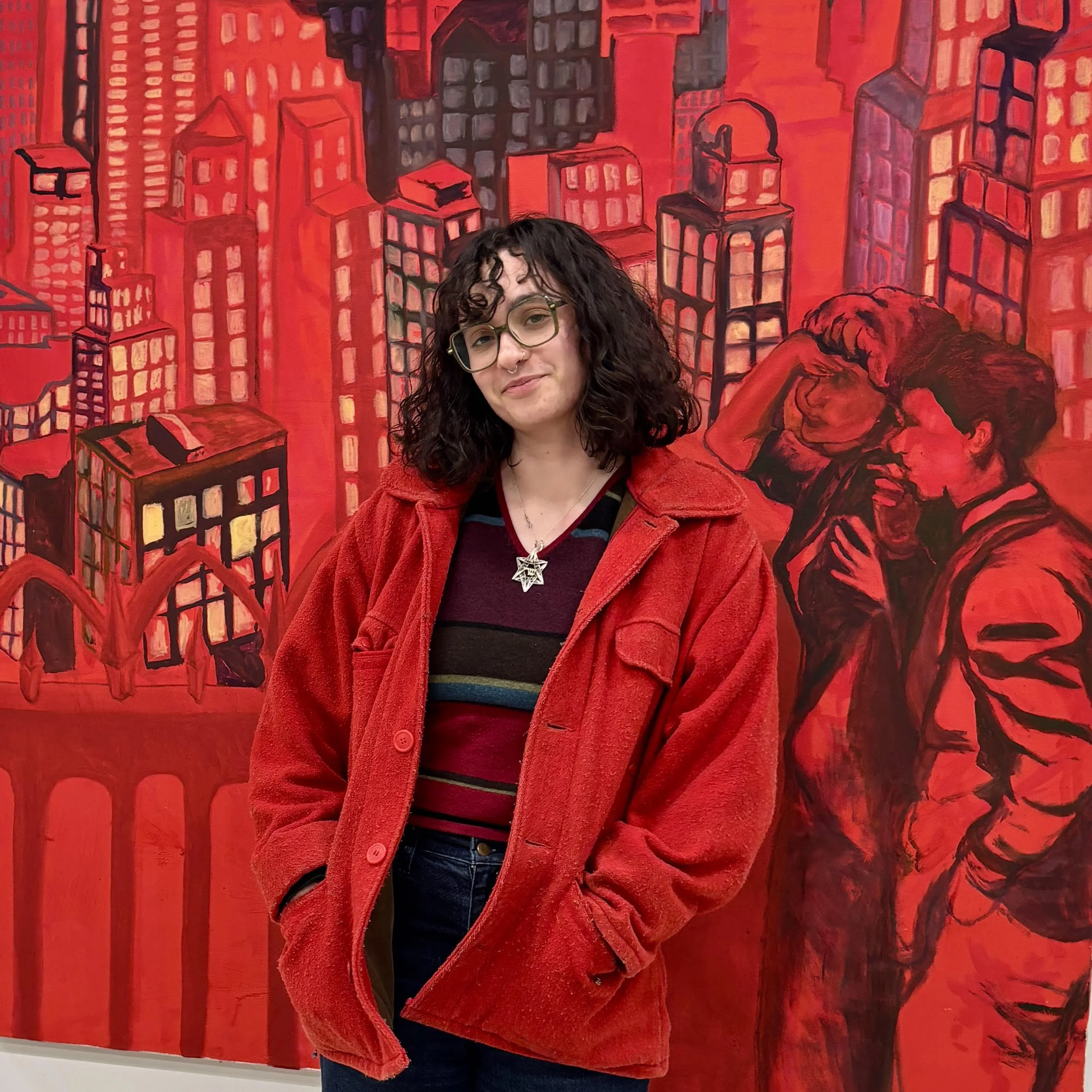 A woman with curly dark hair, glasses, and a star-shaped necklace, standing in front of a vibrant red and black mural depicting city buildings and two people in close conversation.