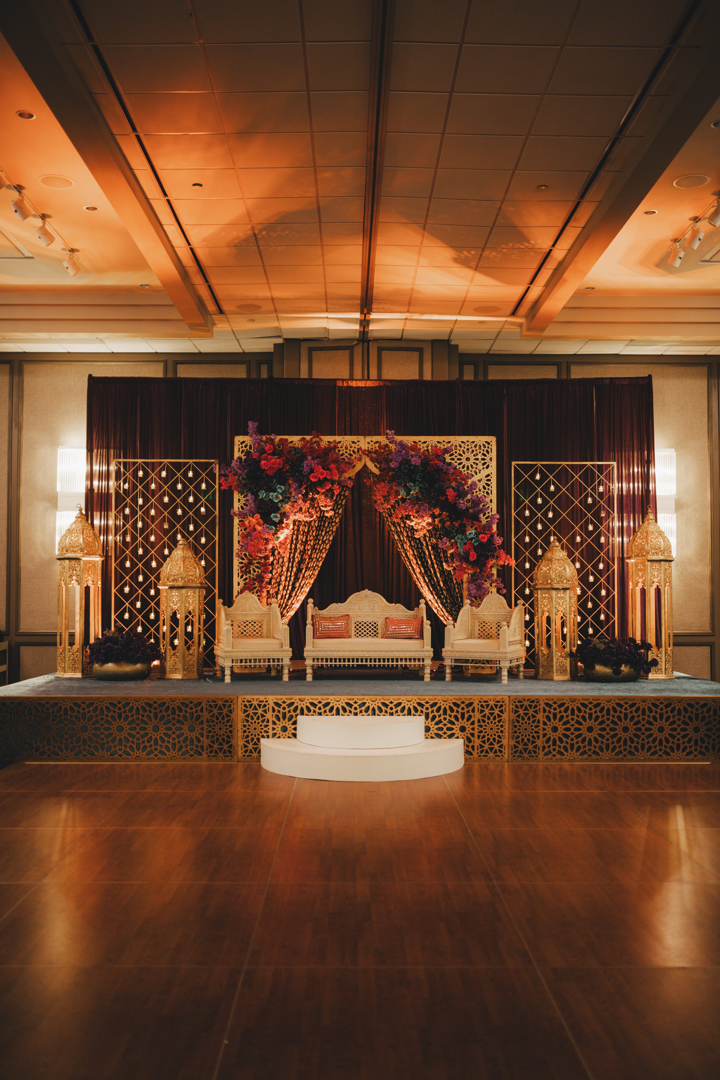 A decorated event stage with ornate white chairs, floral arrangements, gold lanterns, and a backdrop with drapes and lattice panels, set in a banquet hall with wooden flooring.
