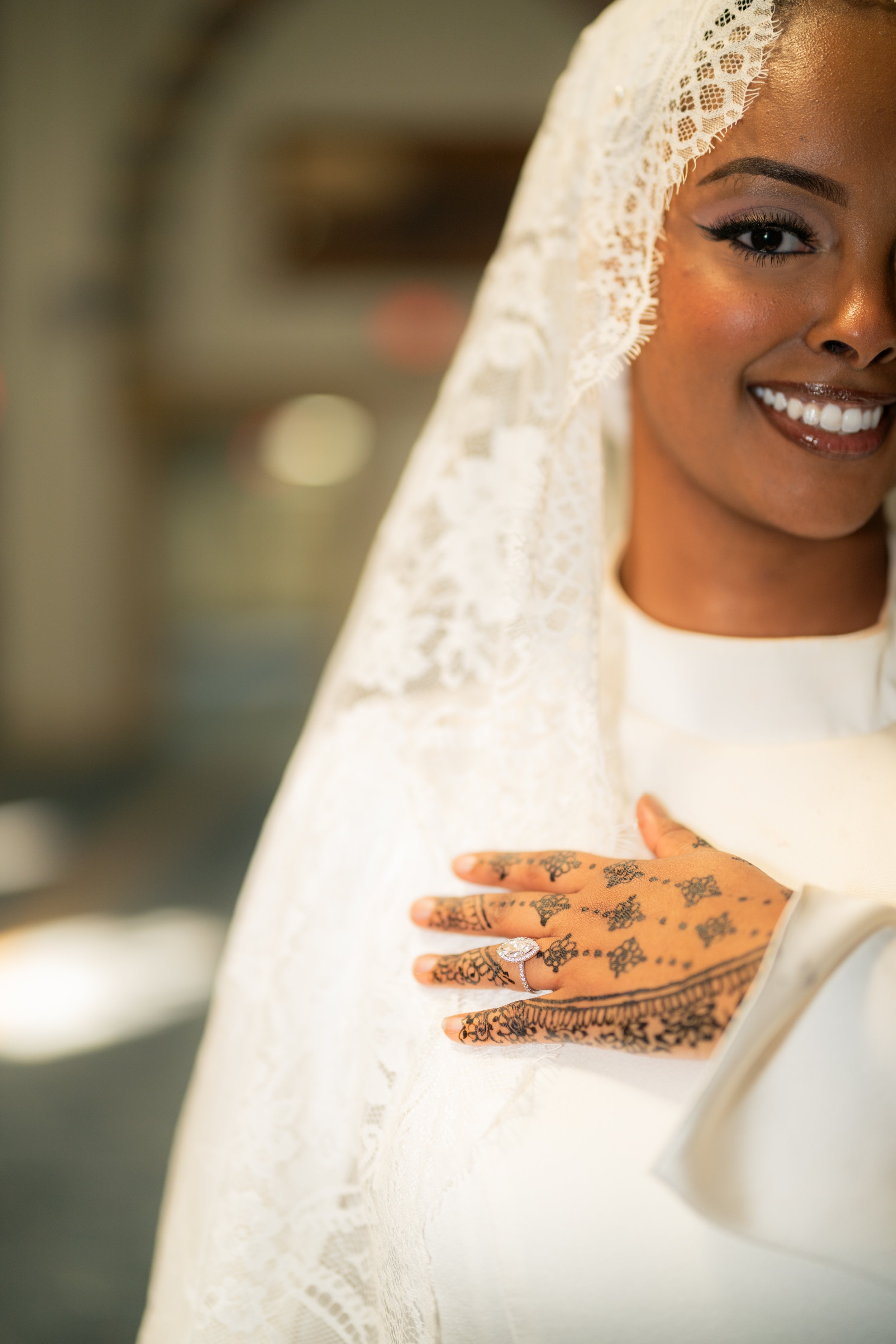 Close-up of a smiling bride with henna-decorated hand on her chest, wearing a lace veil, makeup, and a diamond ring.