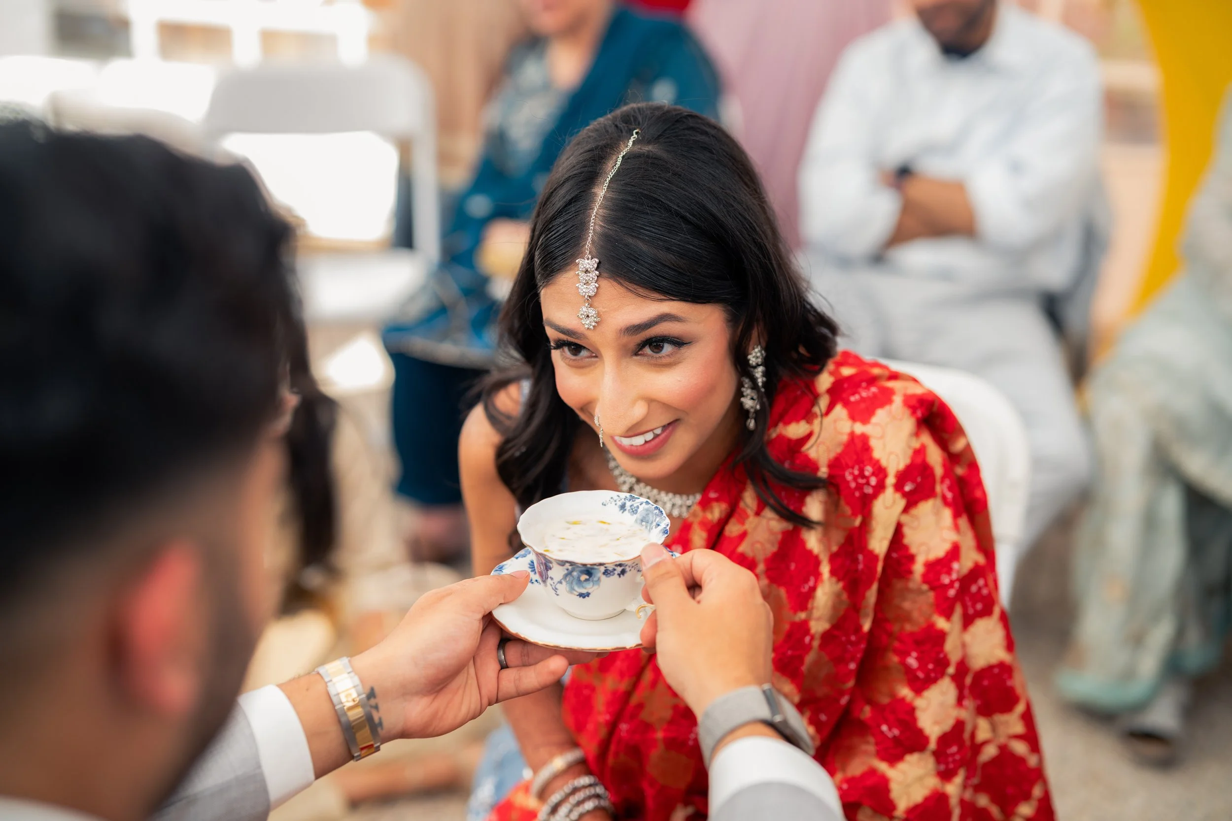 Young woman in traditional Indian attire receiving a sip of tea during a wedding ceremony.