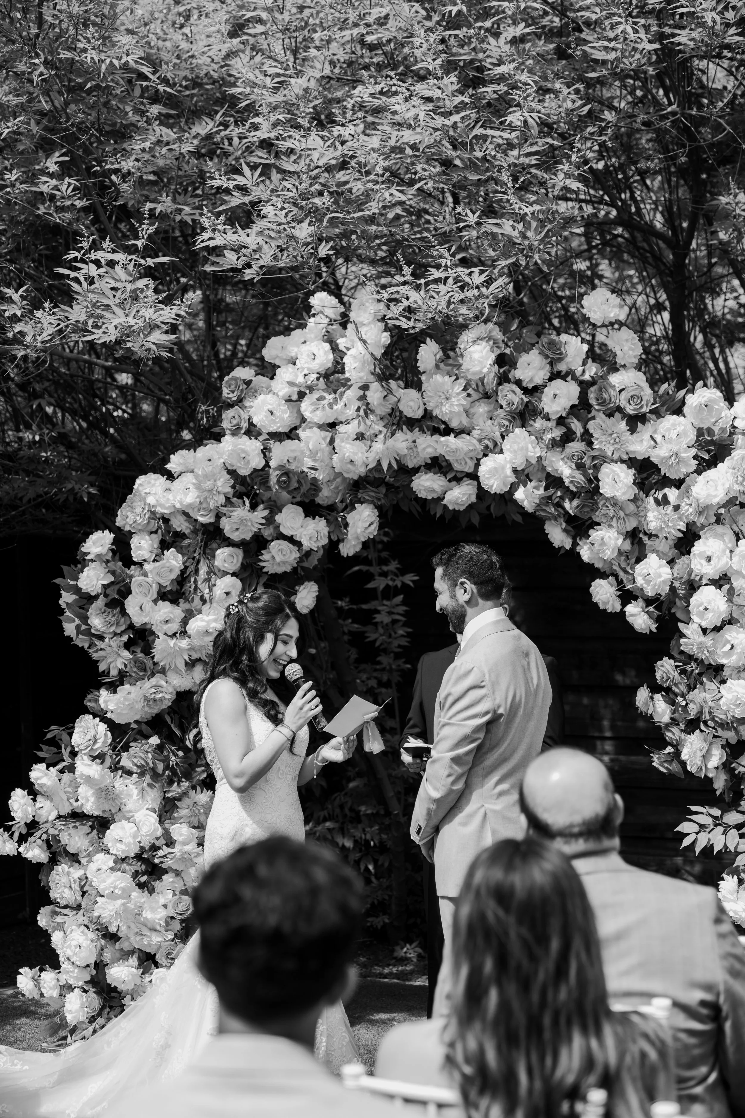 A black and white photo of a wedding ceremony with a bride and groom standing under a floral arch, exchanging vows, with guests seated in front.