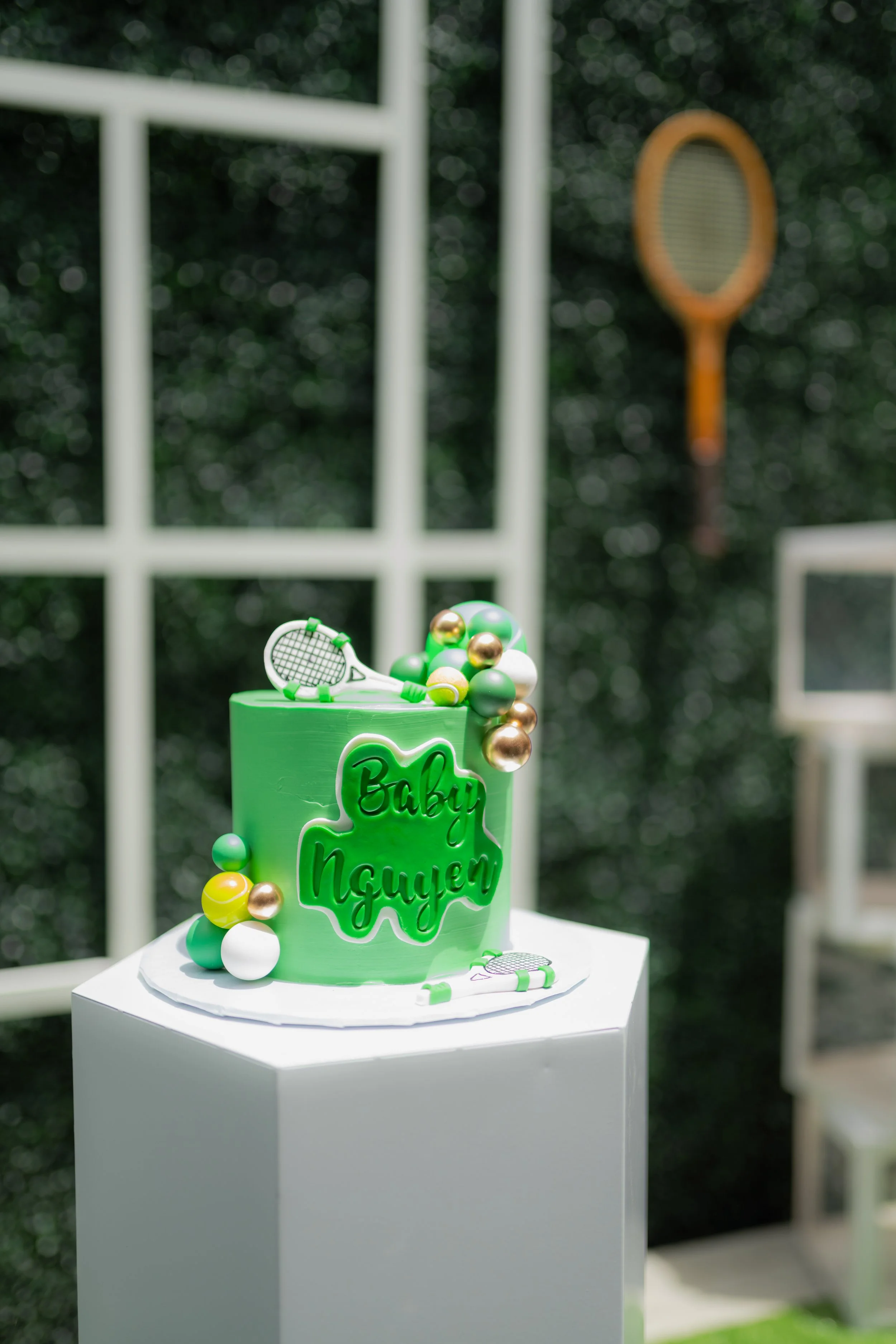 Green cake with tennis-themed decorations, including tennis racket and ball ornaments, with a sign in Cyrillic script, set outdoors on a white pedestal.