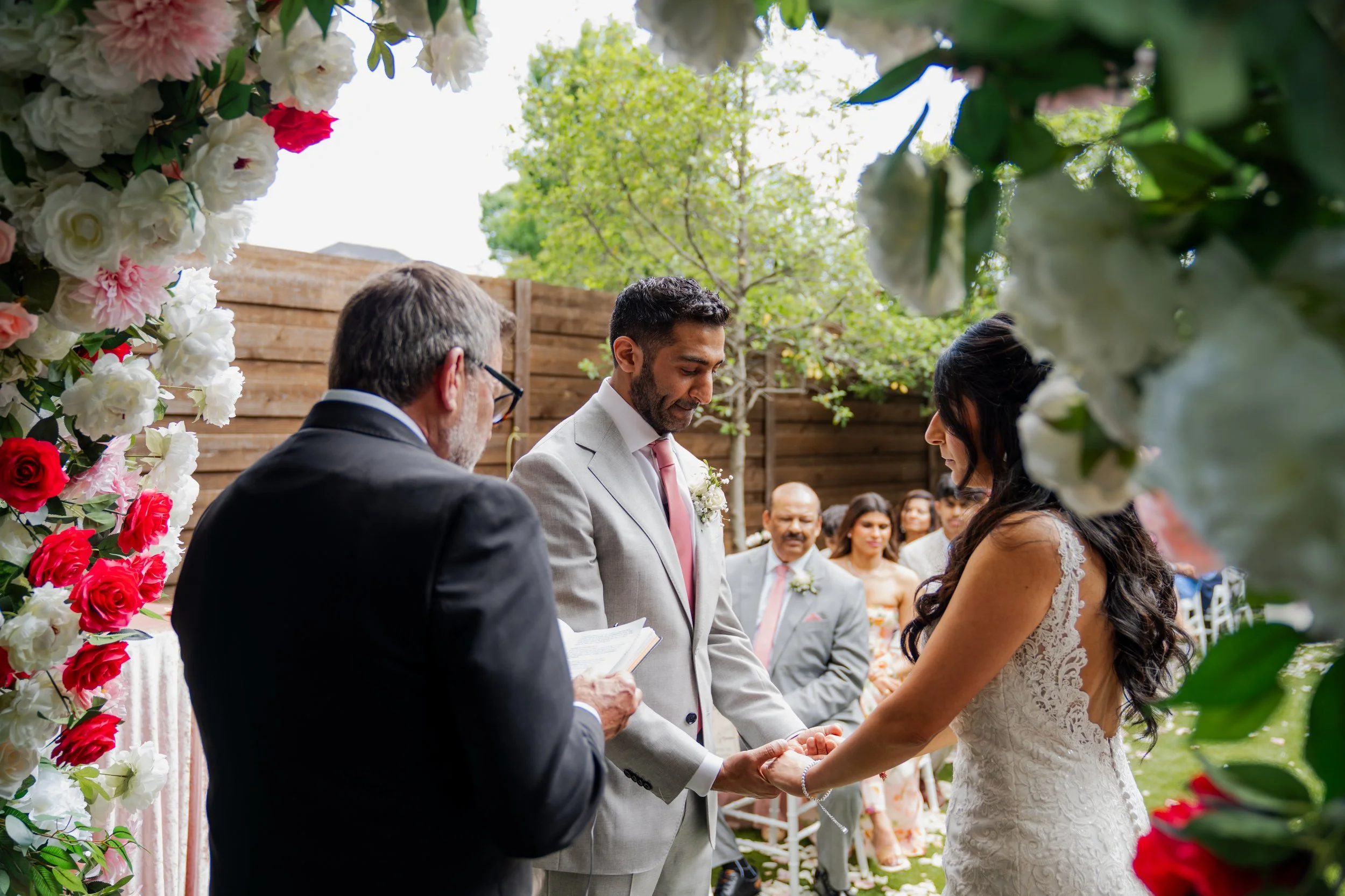A wedding ceremony outdoors with a bride and groom holding hands, exchanging vows, surrounded by floral decorations and guests.