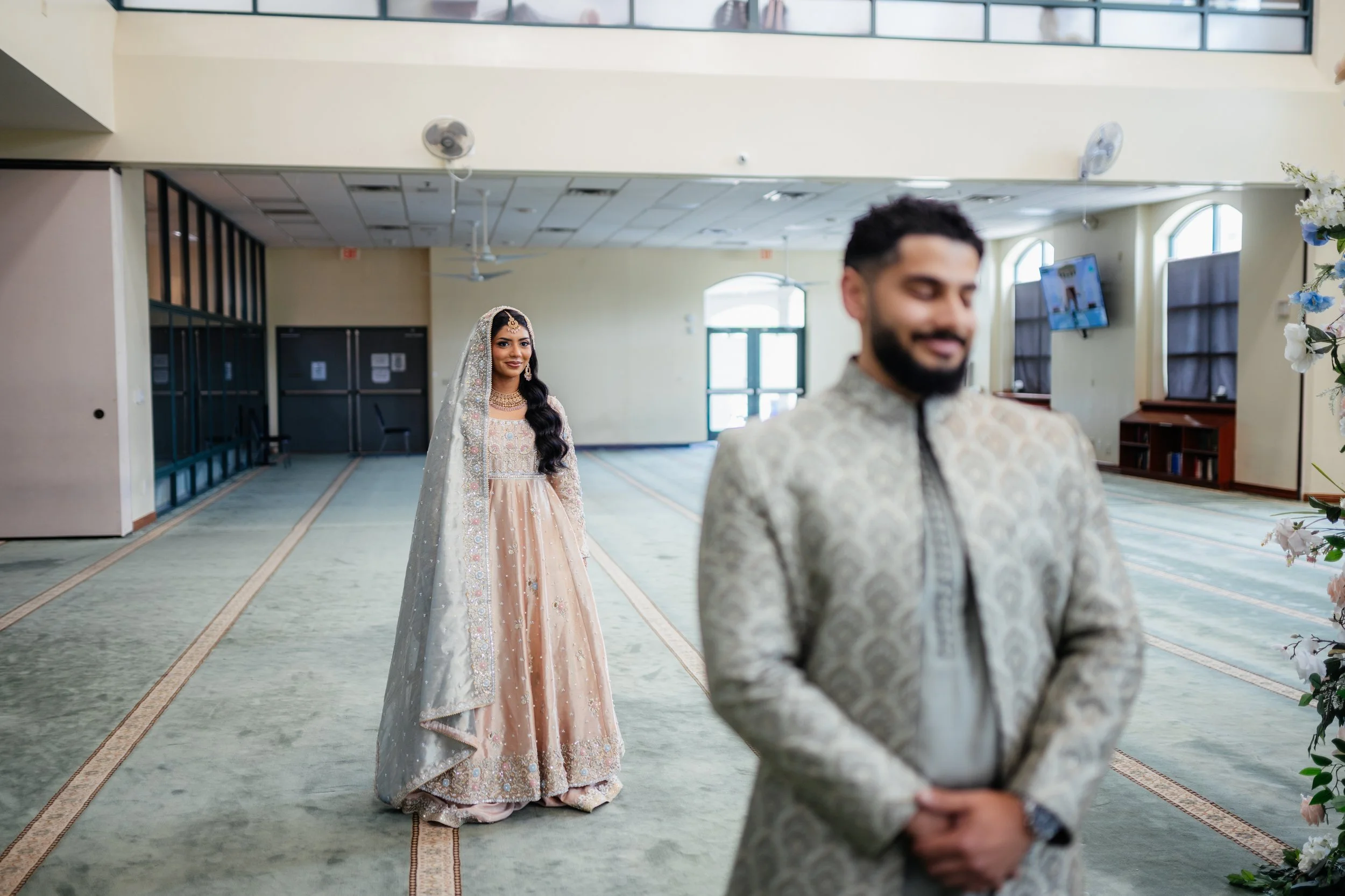 A bride in a traditional beige and gold dress and veil stands in a spacious hall behind a groom in a patterned traditional outfit, who is smiling with his eyes closed and hands clasped. The hall has large windows and a TV screen on the wall.