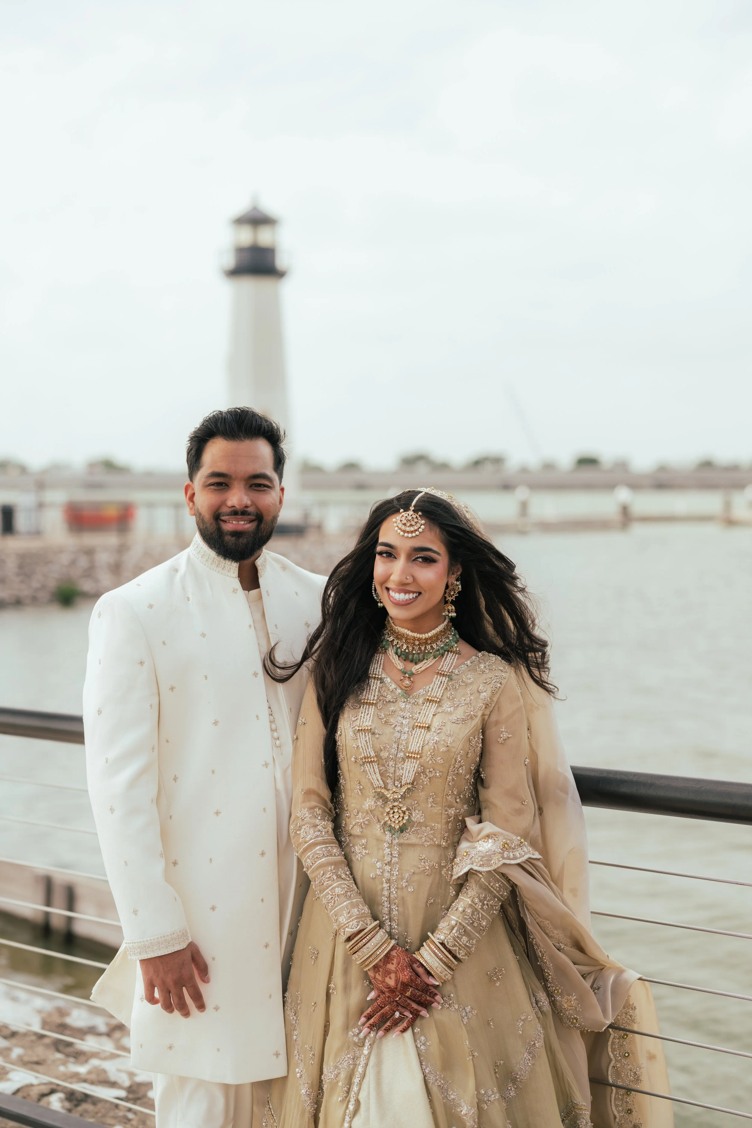 A couple dressed in traditional South Asian wedding attire standing by a river with a lighthouse in the background.