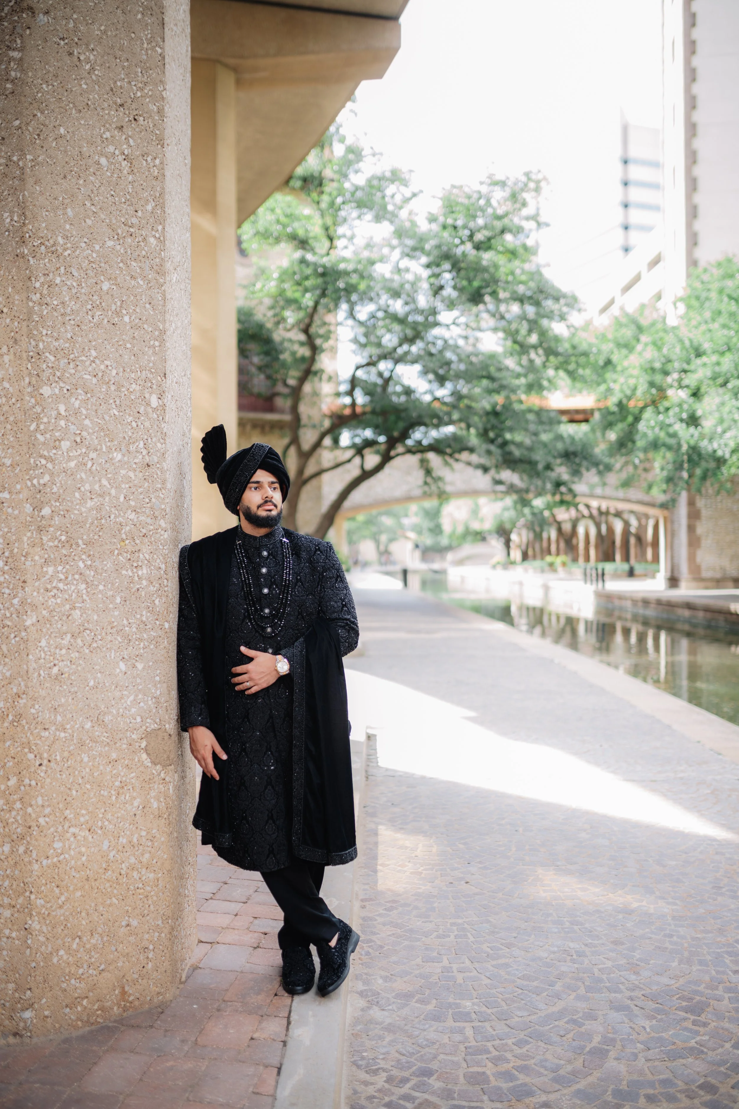 A man wearing a black traditional outfit with a black turban and multiple beaded necklaces, standing against a large beige textured wall with an outdoor cityscape in the background, including trees, a water canal, and buildings.