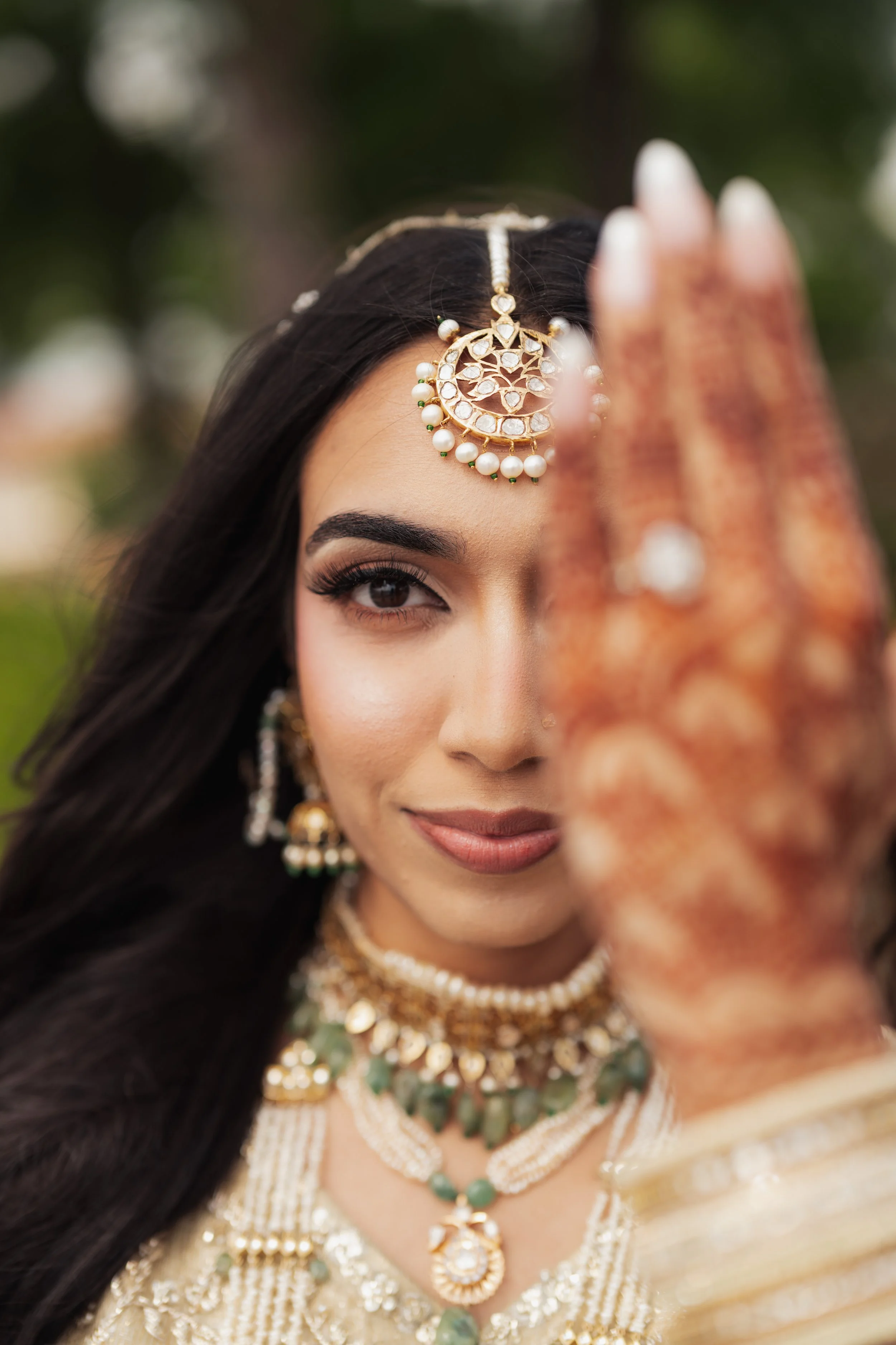 A woman wearing traditional Indian jewelry and attire, with her hand raised in front of her face. Her dark hair and eye are visible, with a blurred green background.