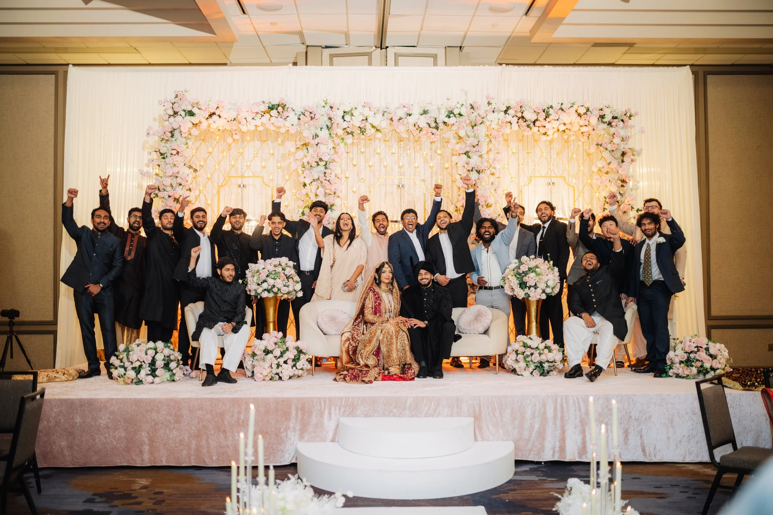 A large wedding reception group photo taken on a stage decorated with pink and white flowers and gold accents. The group consists of men and women, some in traditional and formal attire, with many smiling and raising their fists in celebration.
