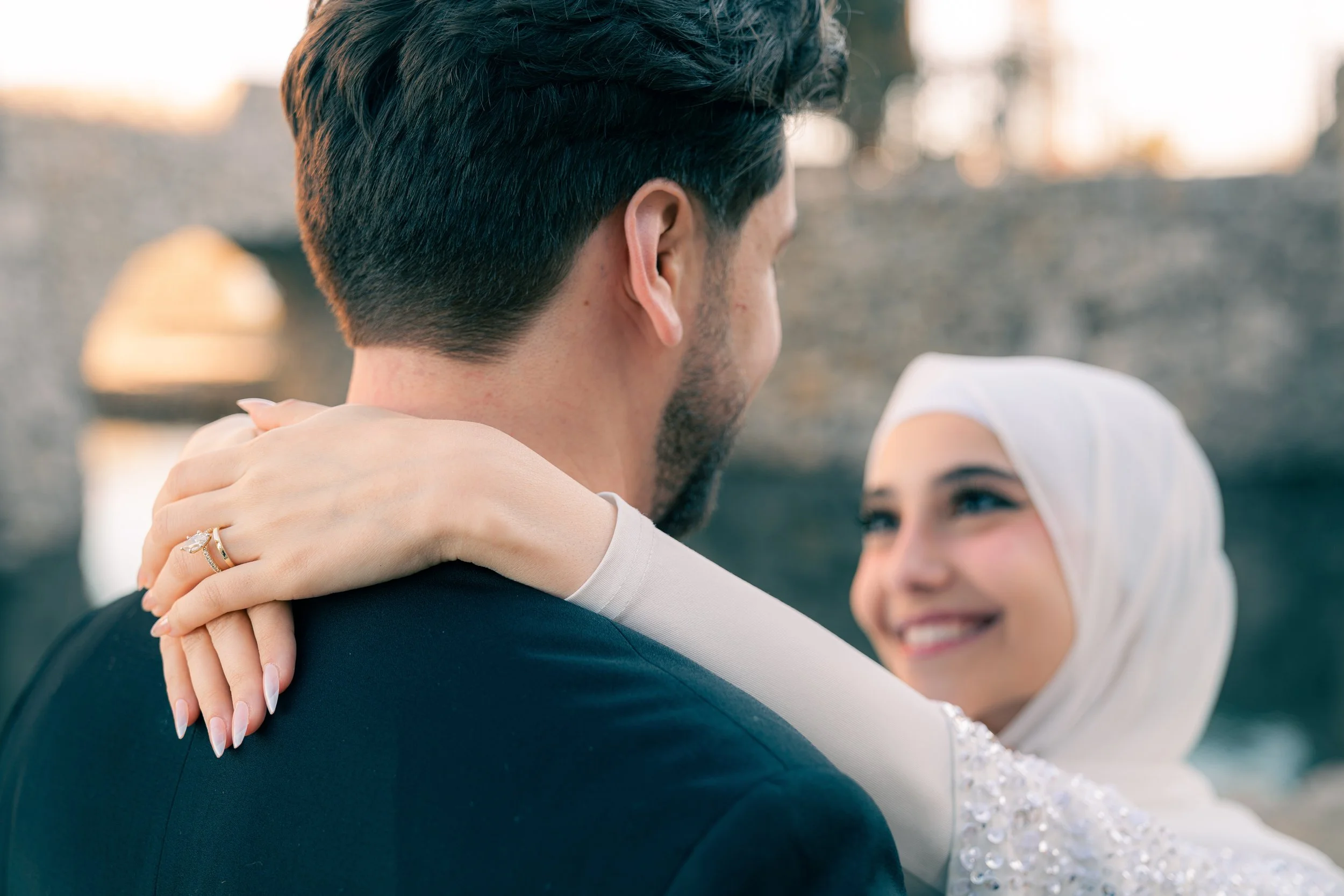 A couple on their wedding day, with the bride wearing a white hijab and the groom in a dark suit, sharing a joyful moment outdoors