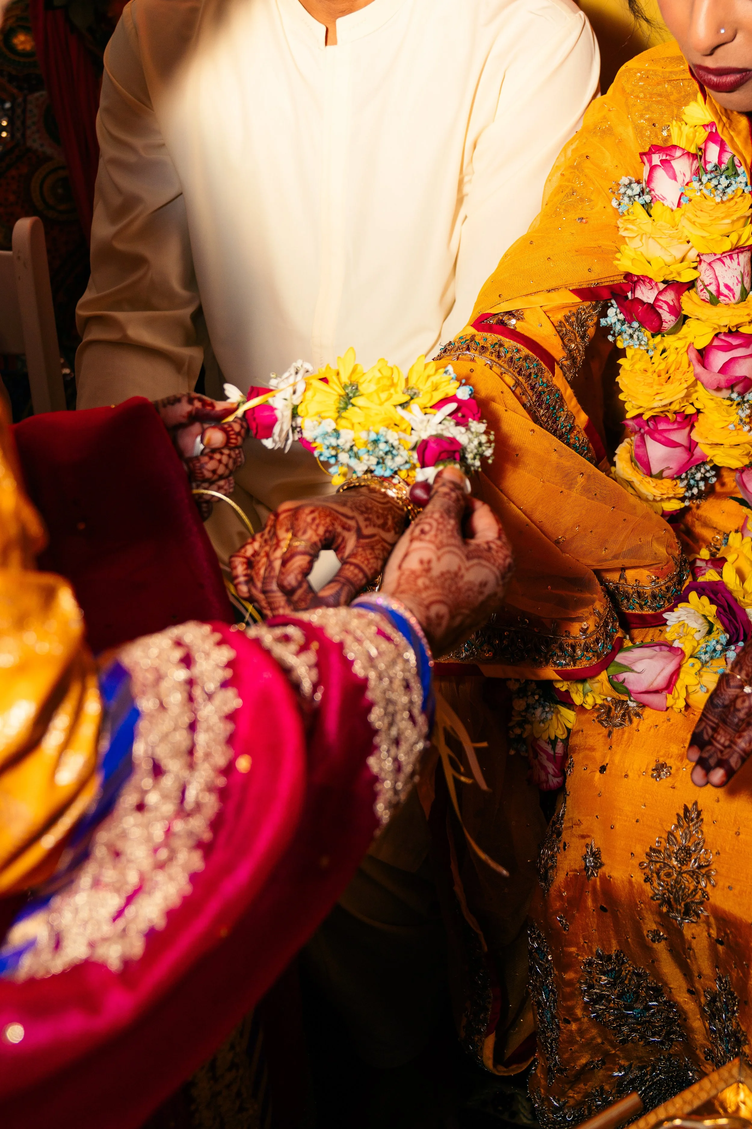 Close-up of a traditional Indian wedding ceremony, showing hands adorned with henna, exchanging flower garlands, with women dressed in colorful, embroidered sarees.