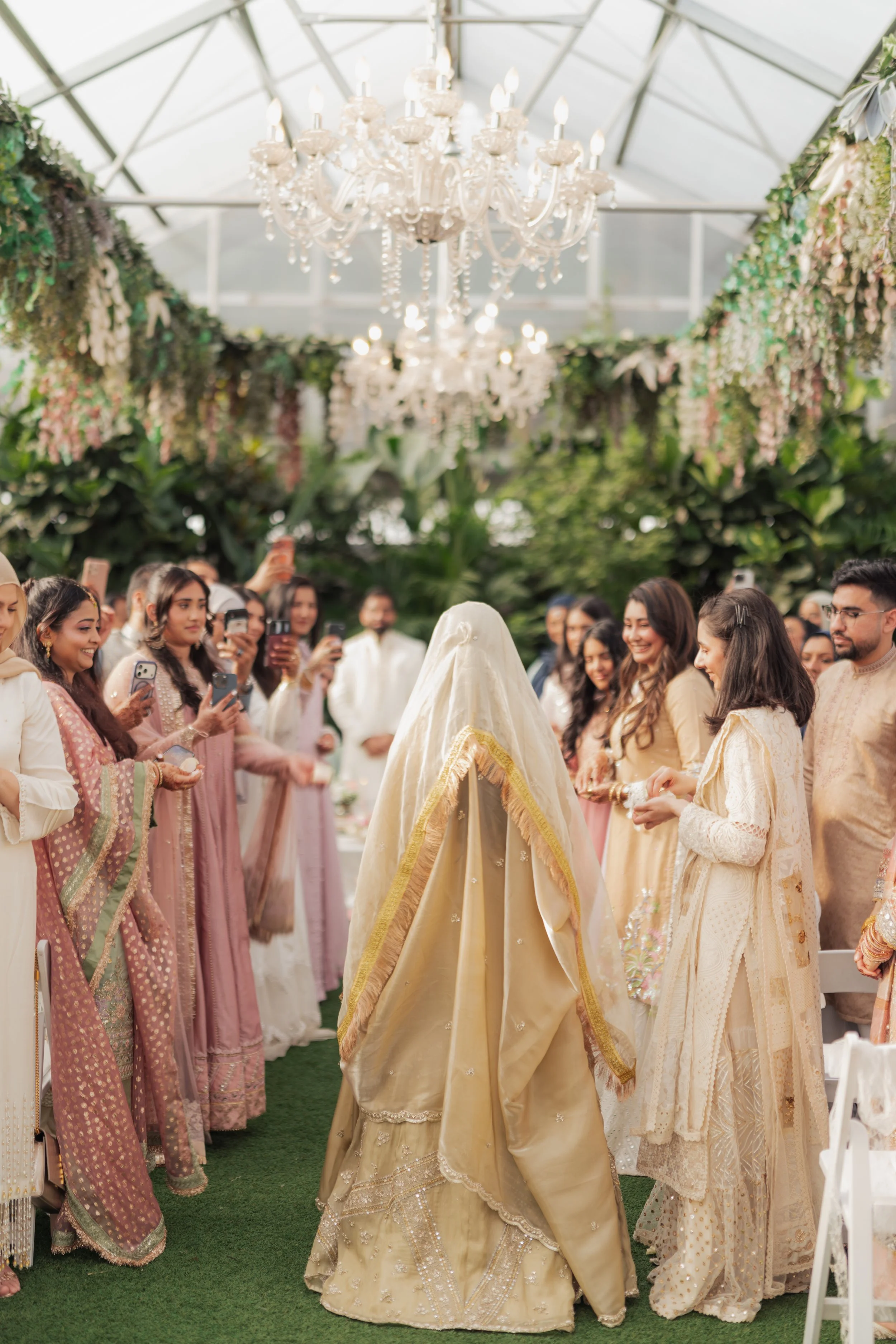 A bride in a gold-colored traditional wedding dress standing among a group of women dressed in pastel-colored traditional attire, with a lush green indoor garden and floral decorations in the background.
