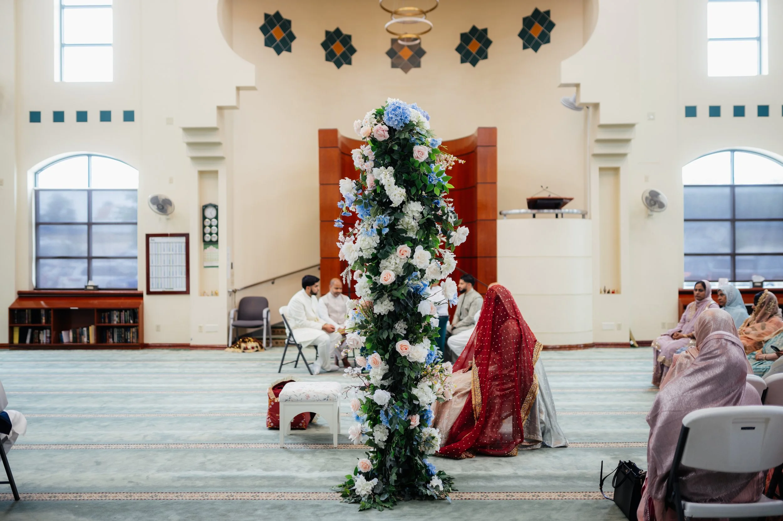 Interior of a mosque decorated for a wedding, with a large floral arrangement in the center, and people seated on the floor, some dressed in traditional attire.