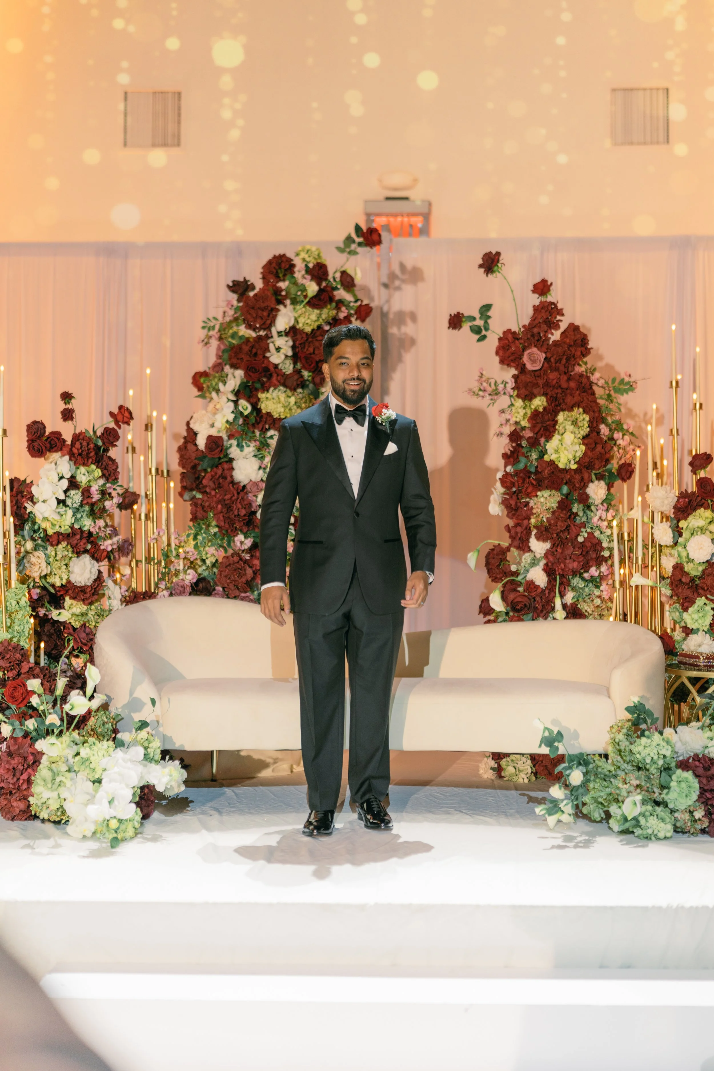 A man in a tuxedo standing in front of a floral wedding backdrop with red, white, and green flowers, lit candles, and a beige sofa.