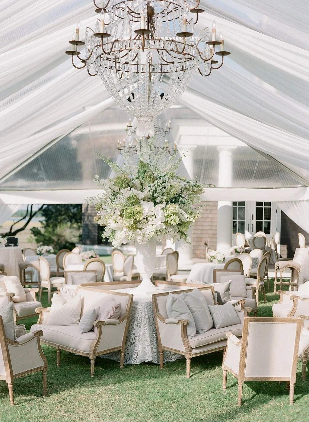 Elegant outdoor event setting under a transparent tent with a large crystal chandelier, floral arrangements, and classic upholstered seating on a grassy lawn.