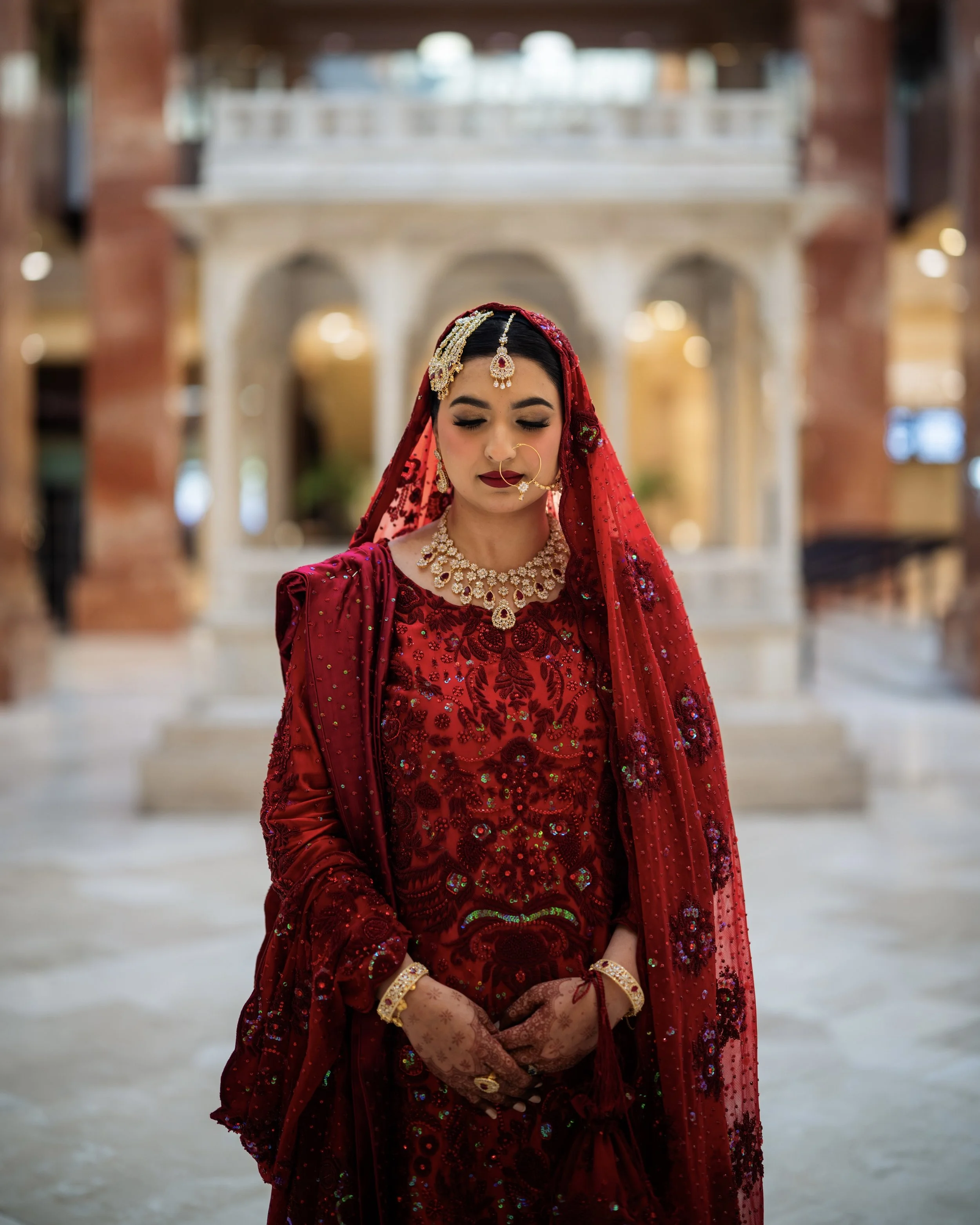 A woman dressed in a red bridal wedding outfit with jewelry, standing indoors with a marble arch and staircase in the background.