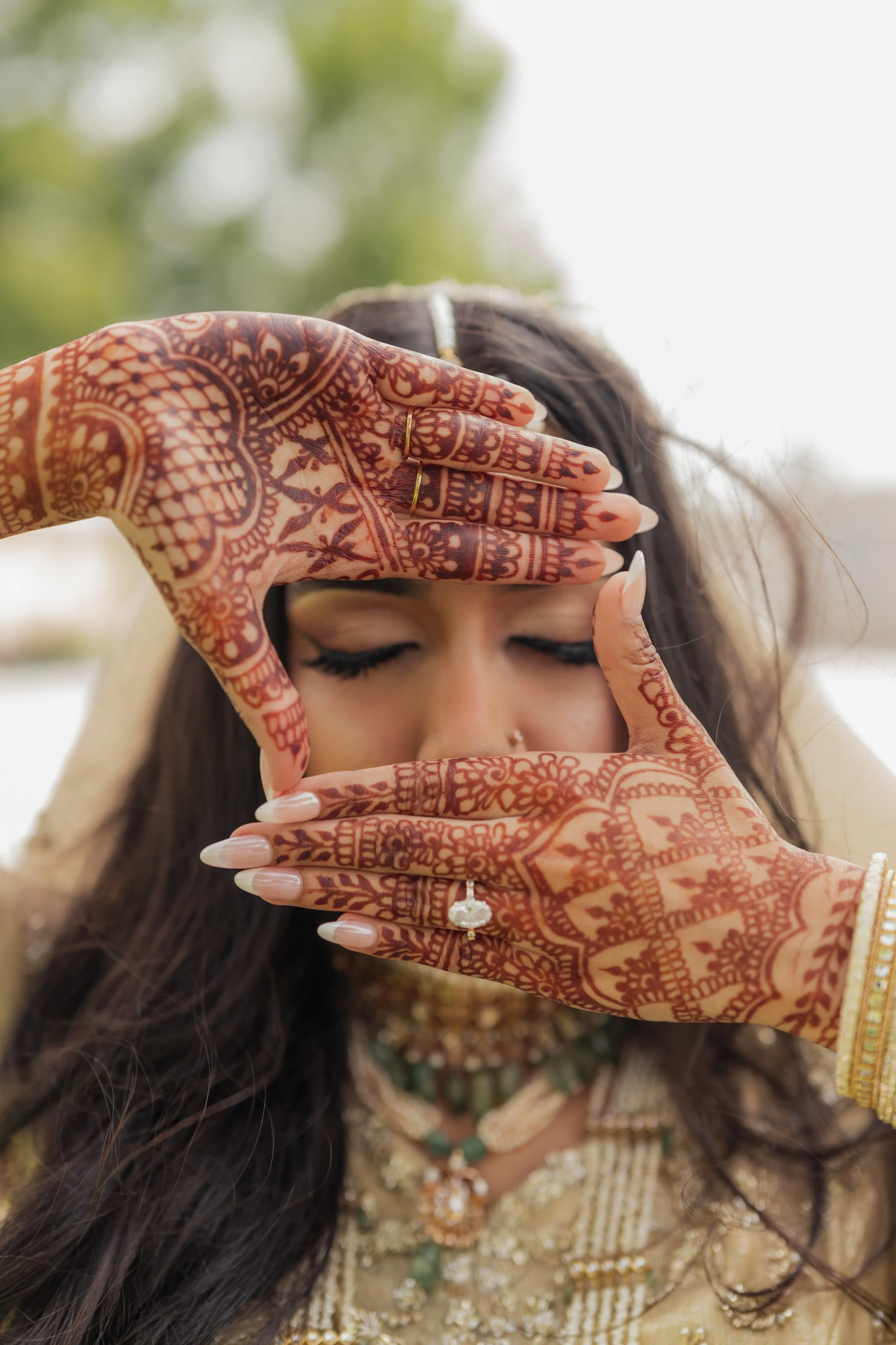 A woman with detailed henna designs on her hands and face, holding her hands in front of her face to frame her eyes. She is wearing jewelry and traditional attire, with a blurred outdoor background.