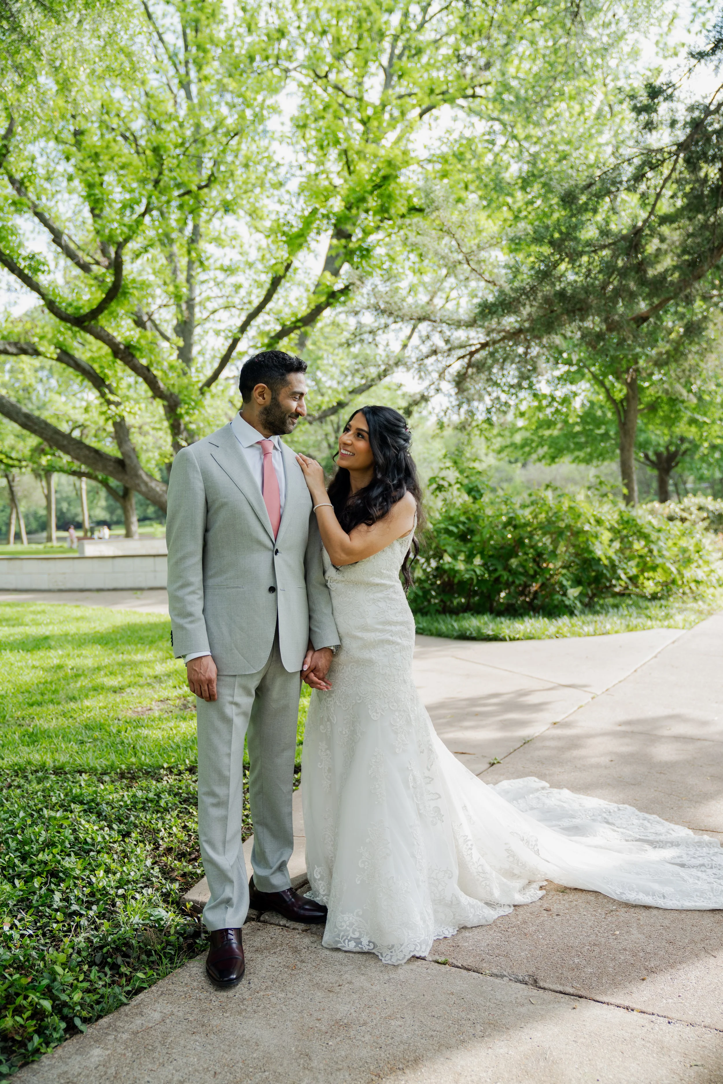 A bride and groom stand close together outdoors in a park with green trees. The groom wears a light gray suit and tie, and the bride wears a white wedding dress with lace details. They look at each other affectionately, holding hands, with the bride 