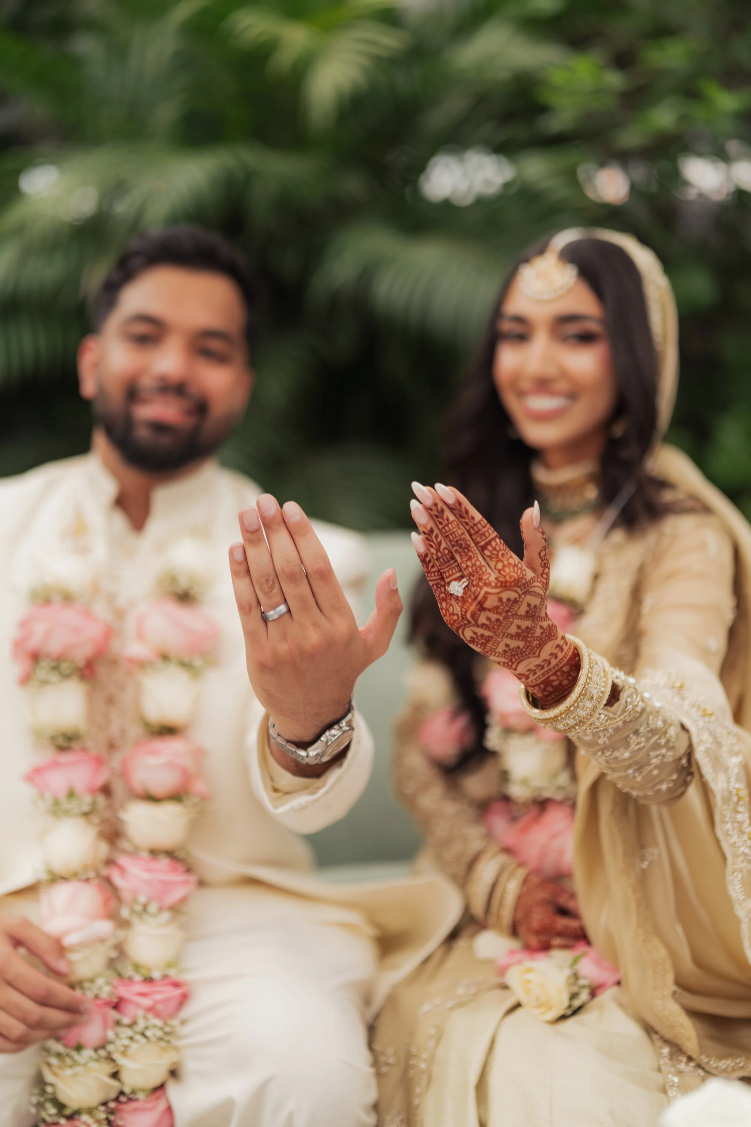 A couple dressed in traditional South Asian wedding attire displaying their wedding rings and hennaed hands during a celebration, with floral garlands and lush greenery in the background.