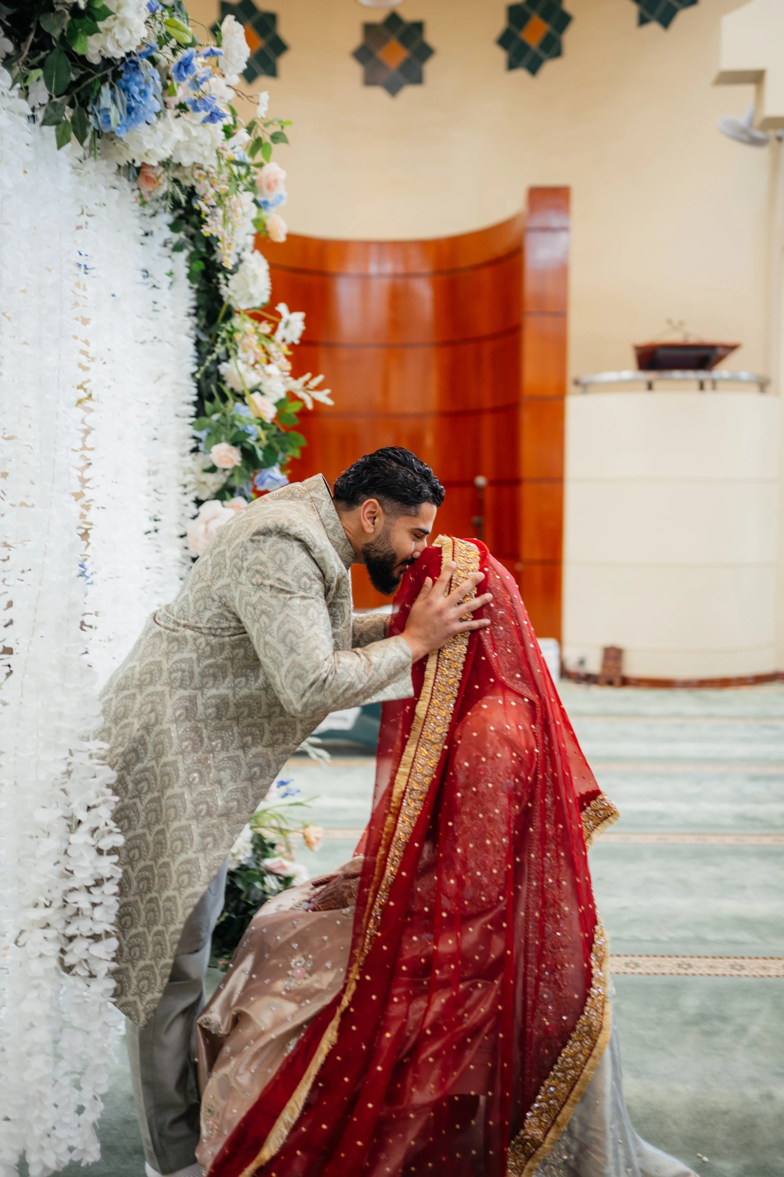 A man dressed in traditional attire leans down to kiss a woman dressed in an elaborate red and gold wedding dress during a wedding ceremony.