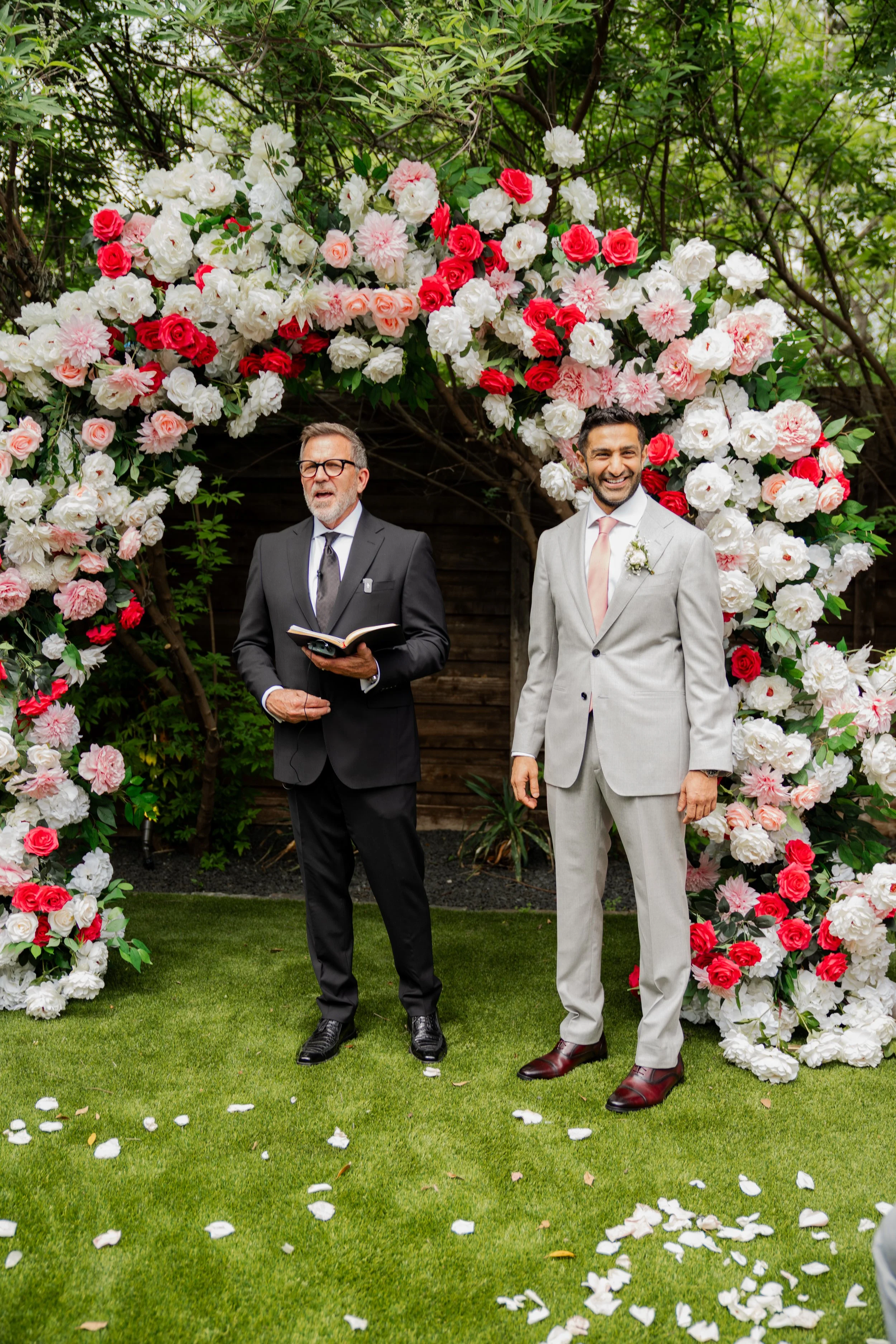 Two men in formal attire standing under a floral arch at a wedding ceremony outdoors. One man is officiating, holding a book, while the other is smiling. Flower petals are scattered on the grass in front of them.