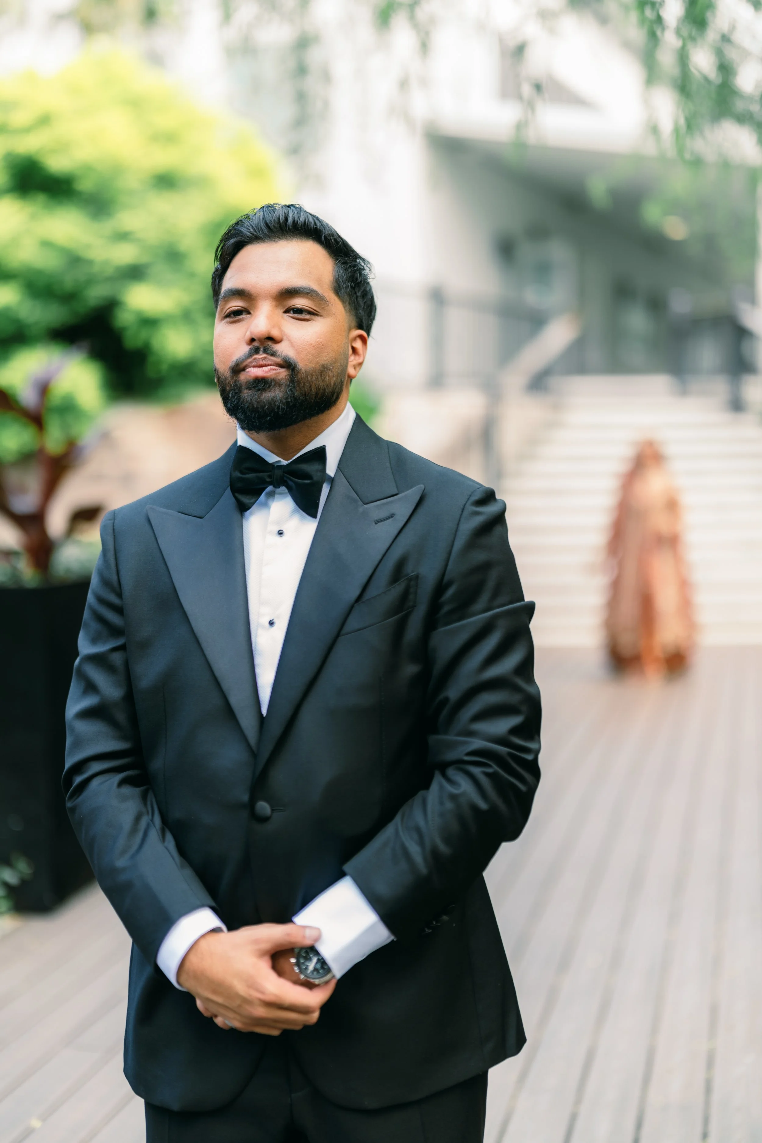 A man in a tuxedo standing outdoors with greenery and a house in the background. He has dark hair and beard, holding a piece of paper and a watch visible on his wrist.