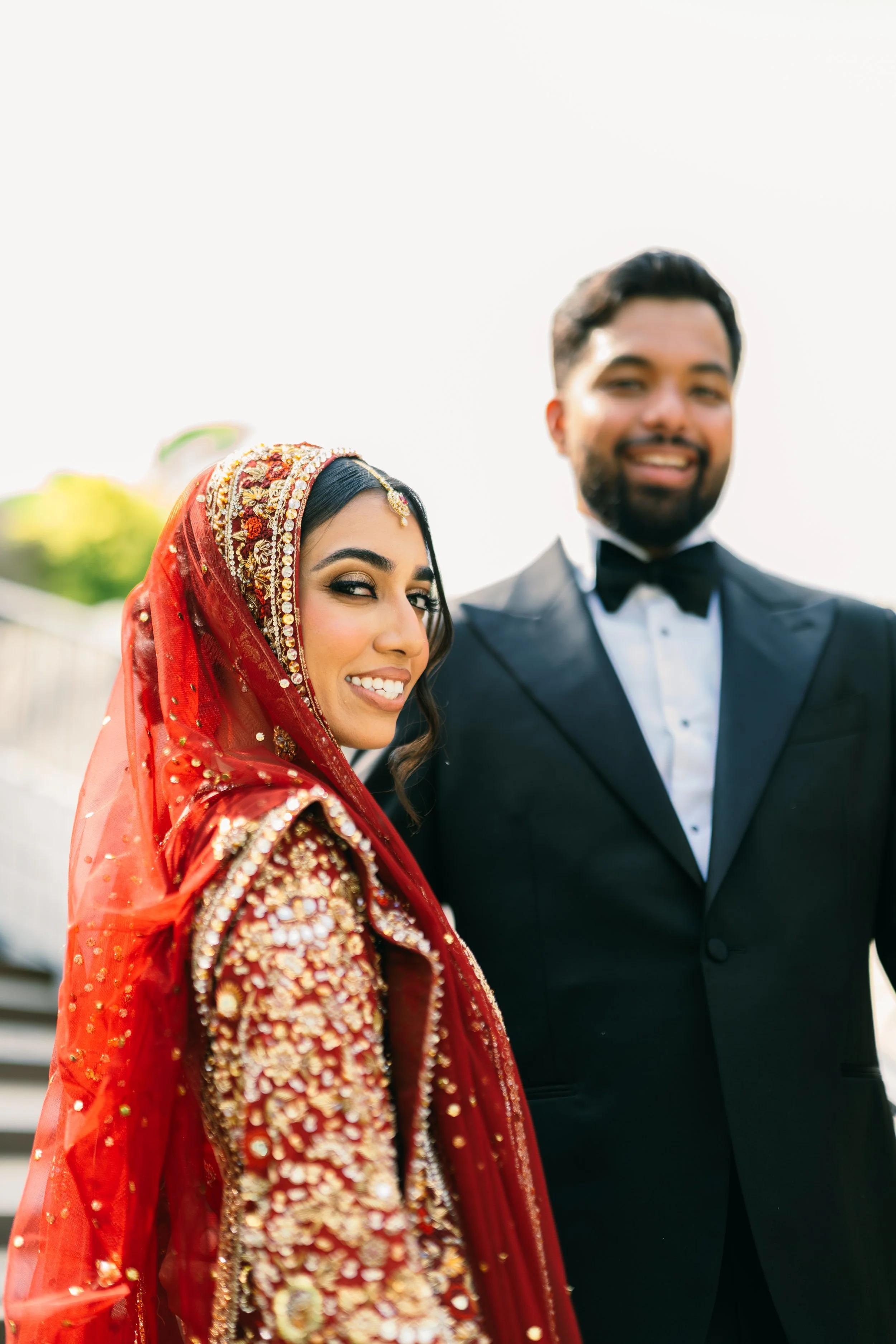 A smiling woman dressed in traditional red South Asian wedding attire with intricate embroidery and jewelry next to a man in a black tuxedo and bow tie, both outdoors with a bright sky in the background.