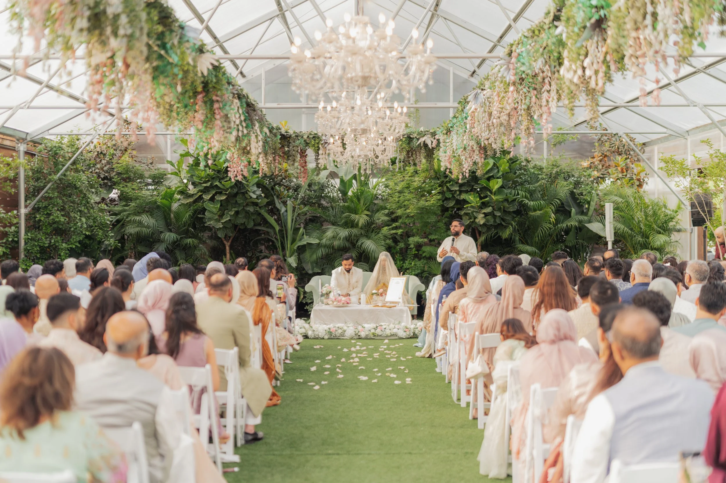 Wedding ceremony inside a glass greenhouse with floral and chandelier decorations, people seated facing the couple.