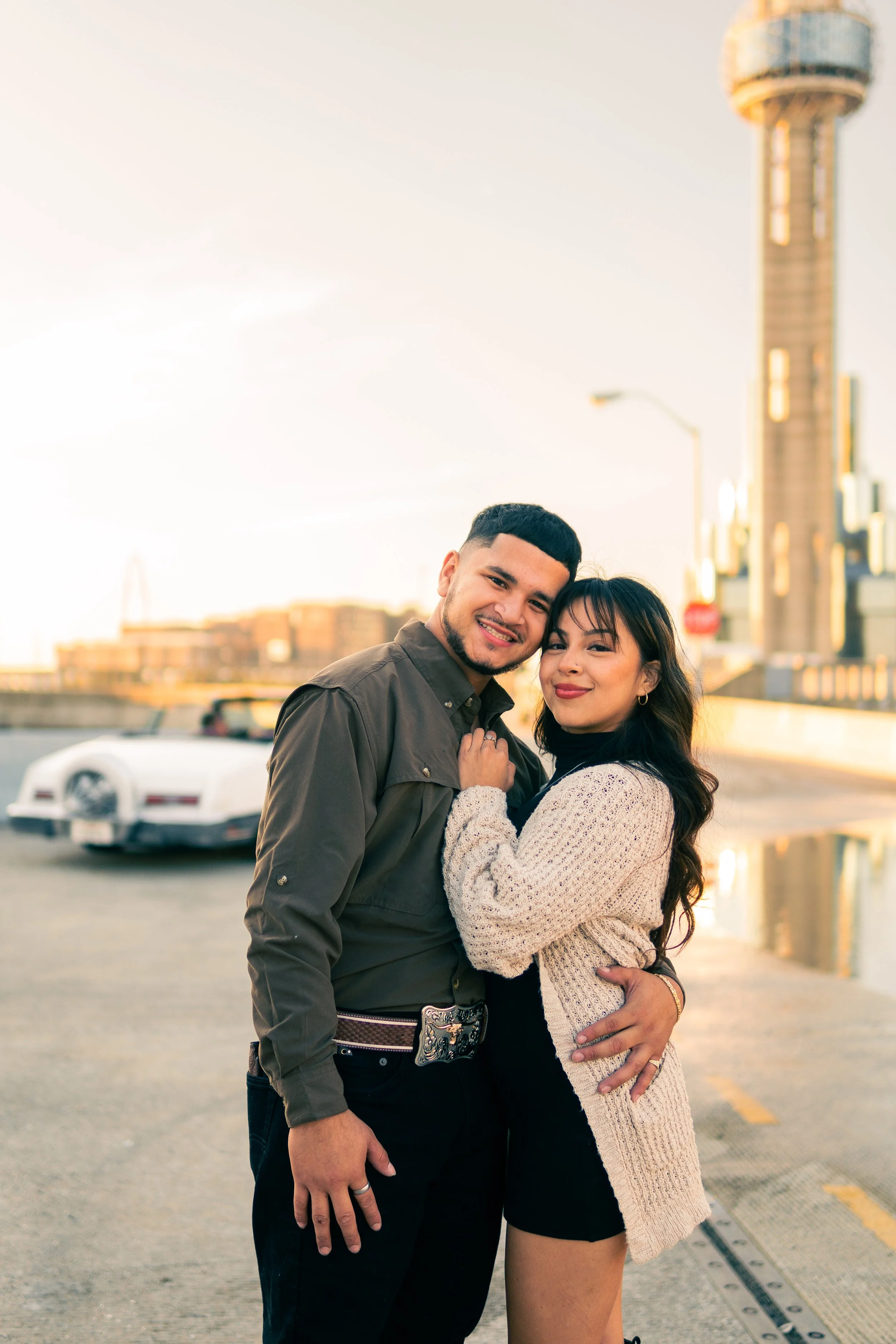 A smiling couple hugging outdoors near a city skyline at sunset, with a white car in the background.
