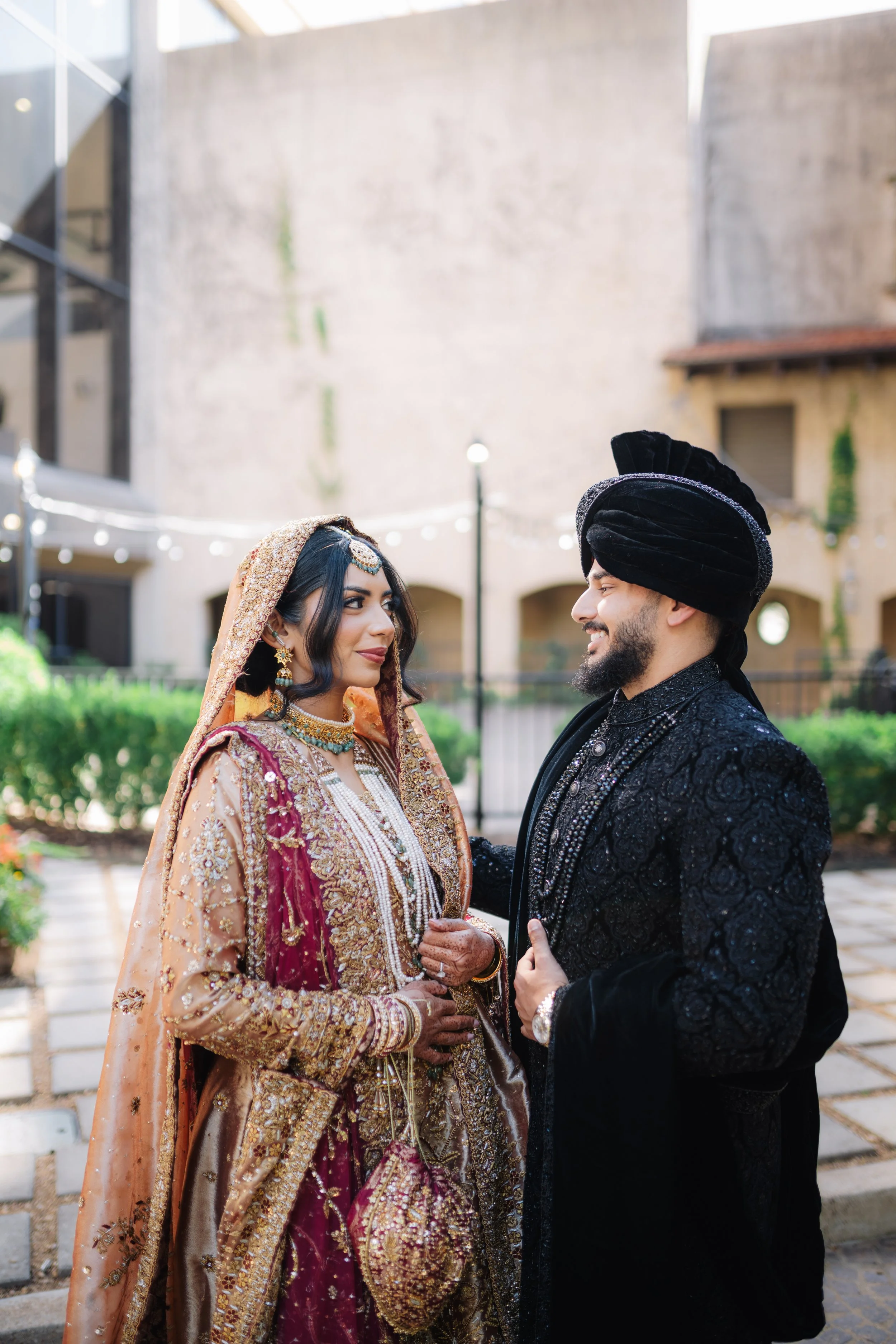 A South Asian bride and groom in traditional attire standing outdoors, facing each other and smiling.