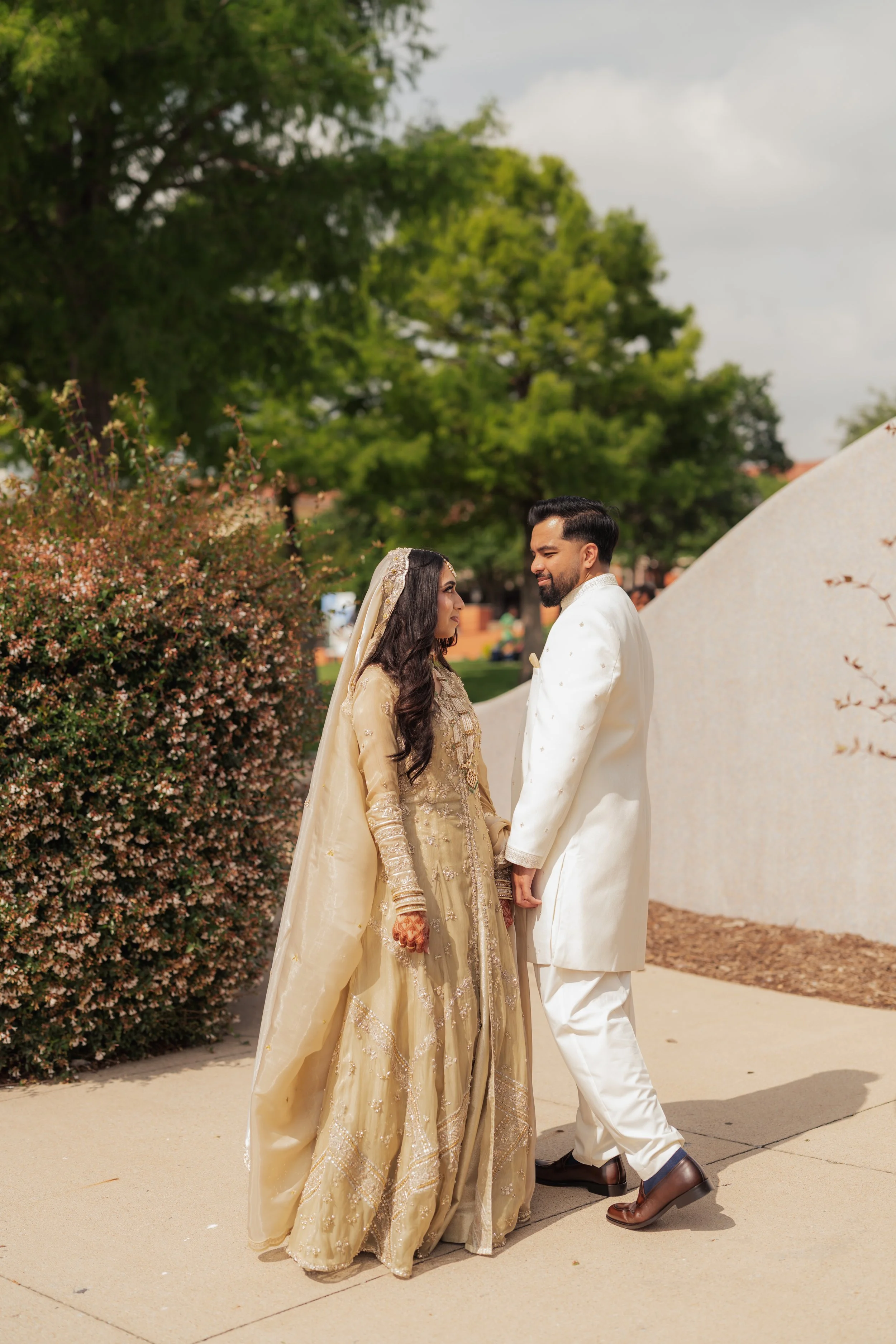A man and woman dressed in traditional South Asian wedding attire standing outdoors, holding hands and looking at each other, with greenery and a cloudy sky in the background.