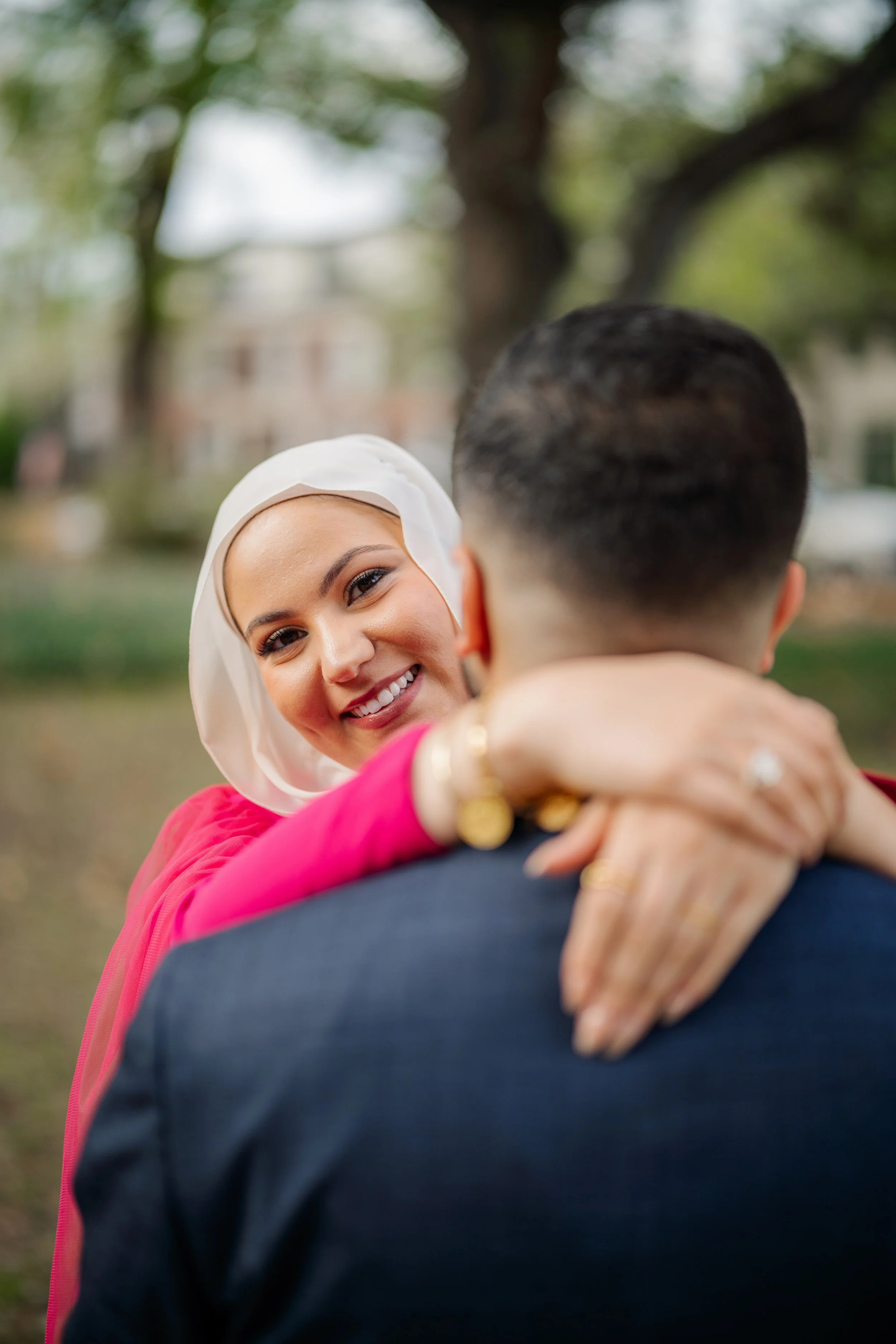 Woman with a white headscarf smiling and hugging a man in a suit outdoors with trees in the background.