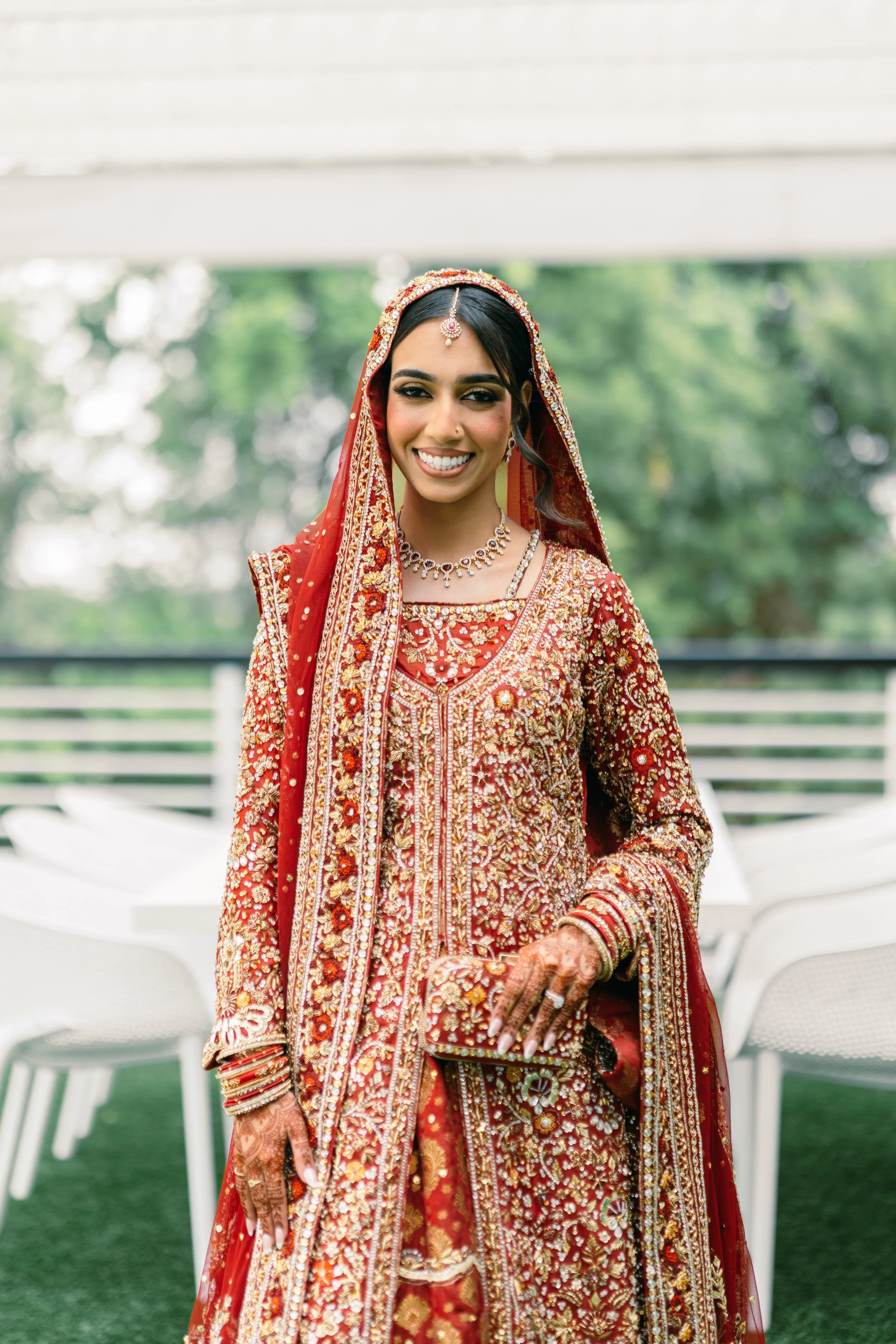 A smiling woman dressed in red and gold traditional South Asian wedding attire, jewelry, and henna, standing outdoors with greenery and white chairs in the background.