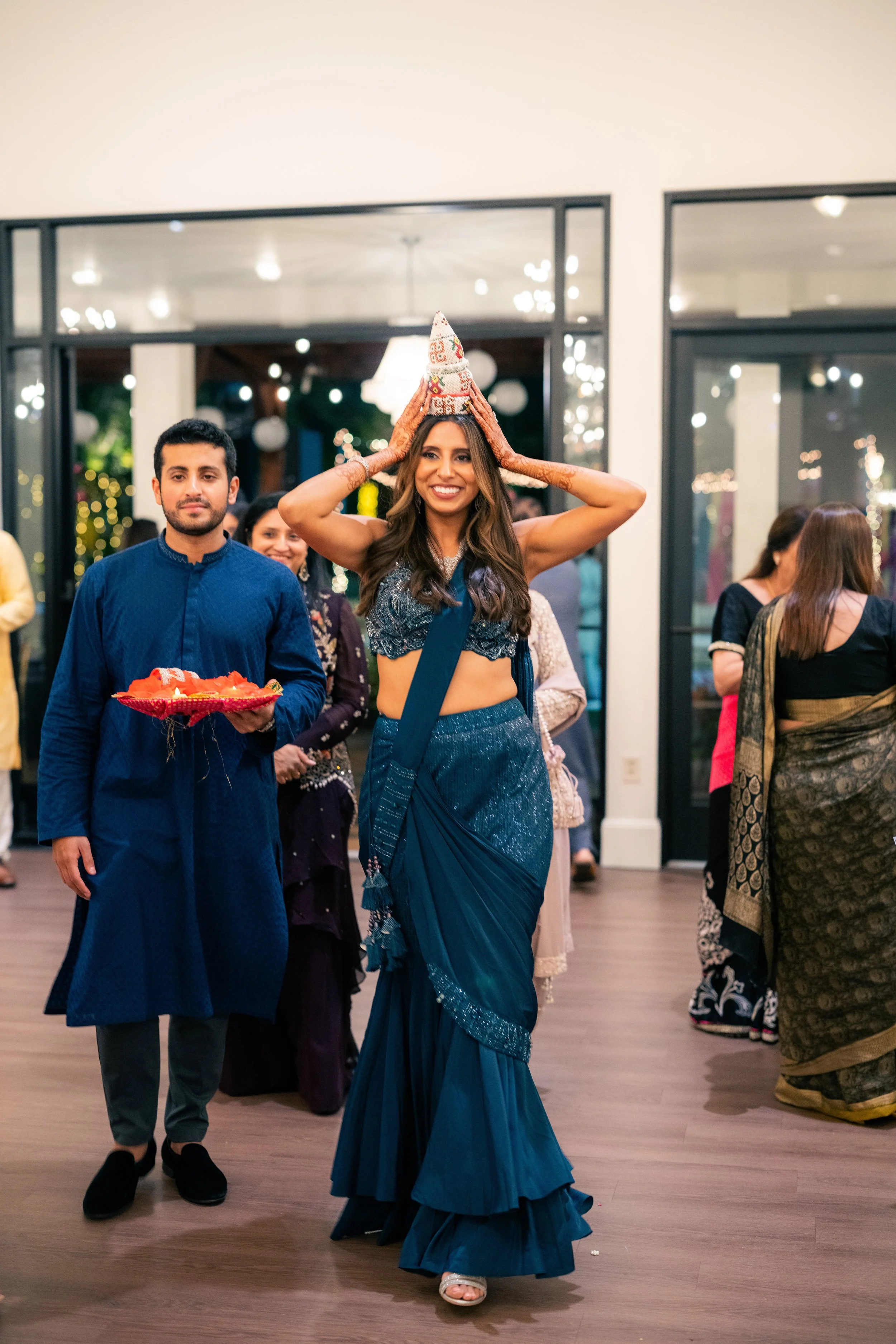 A woman in a blue saree is smiling and placing a traditional headdress on her head during a celebration. She is surrounded by other people in traditional attire, and the setting appears to be indoors at a festive event.